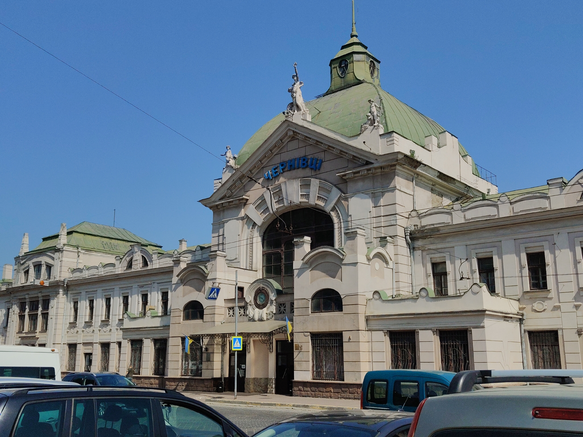 The image shows the Chernivtsi Railway Station in Chernivtsi, Ukraine, on a clear, sunny day. The station building, with its distinctive green dome and ornate classical architecture, is the central focus. The facade is light-colored, with prominent arched windows and decorative sculptures adorning the roofline. Blue lettering, spelling out "ЧЕРНІВЦІ" (Chernivtsi), is visible above the main entrance. Ukrainian flags are displayed near the entrance. In the foreground, several vehicles, including a white van and various cars, are parked or driving on the street in front of the station. The sky is a vibrant, cloudless blue, and overhead power lines are visible. The scene depicts a bustling transportation hub with a historical architectural backdrop.
