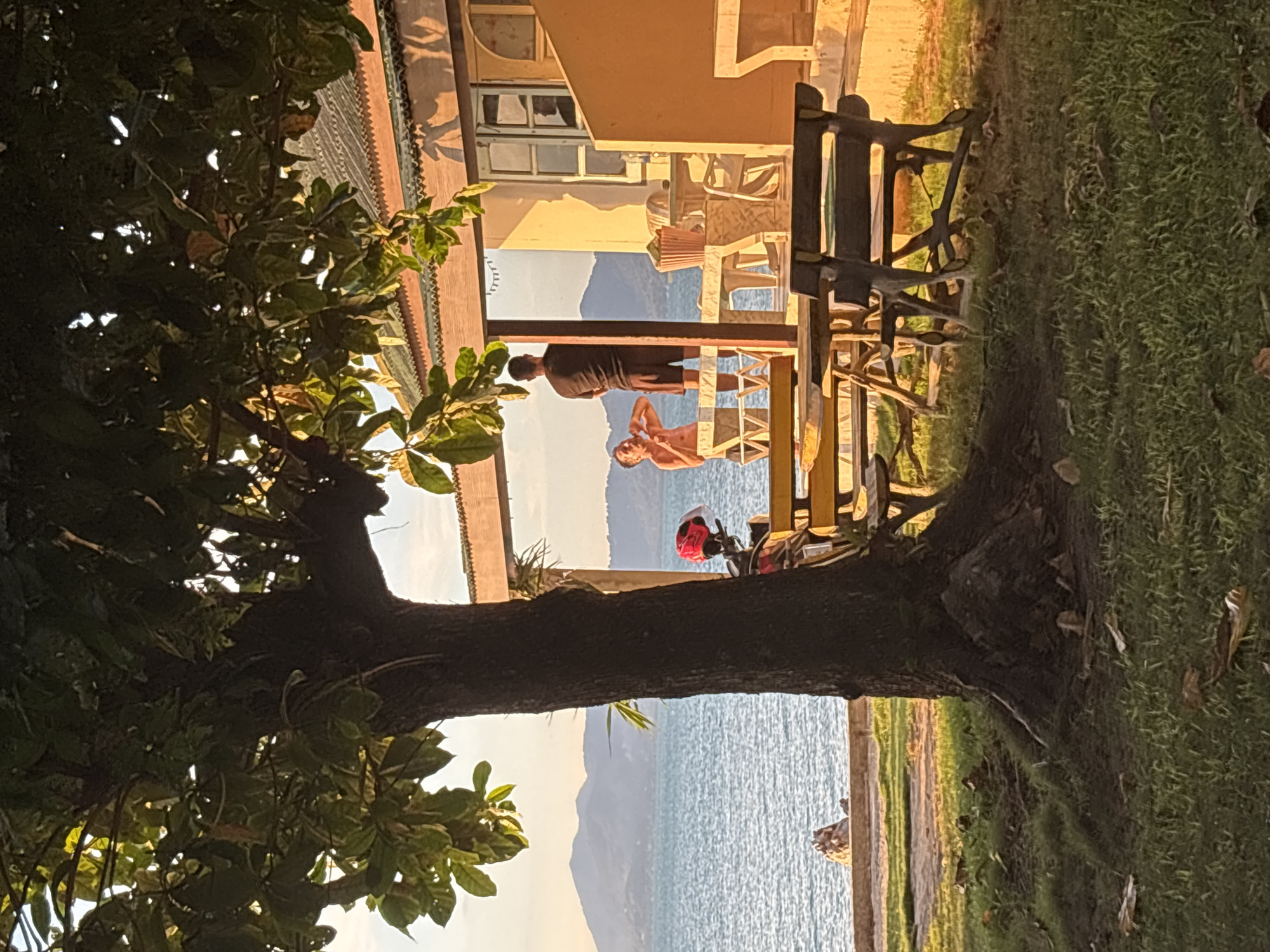Two people stand on a porch, visible through a tree trunk and branches in the foreground. The porch is attached to a tan-colored building with a window. Behind them, the ocean meets a distant mountain range under a bright sky. A red motorcycle helmet sits on a table near the people. There is a bench on the right of the image. Green grass is seen along the bottom right. The location is in Baan Bo Phut, Thailand.