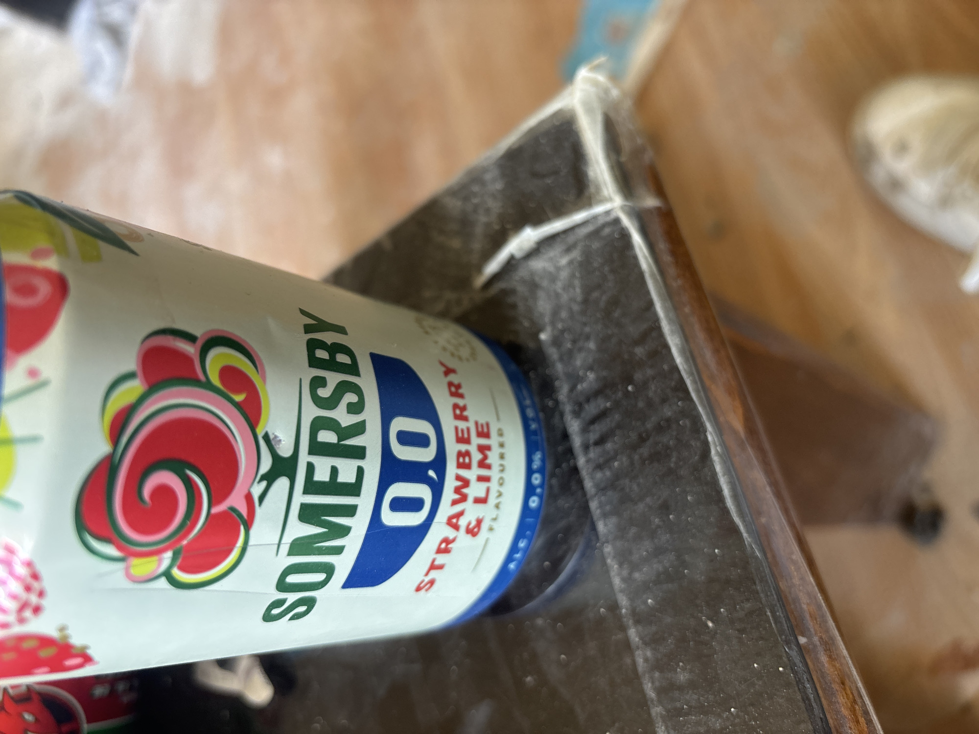 A close-up, angled shot shows a can of Somersby 0.0 Strawberry & Lime flavored cider. The can is light green with red and pink floral graphics and green lettering. The word "SOMERSBY" is prominently displayed in green text, followed by "0.0" in a blue circle. Below that, "STRAWBERRY & LIME" is written in red text. At the bottom, "ALC. 0.0%" is visible in small print. The can rests on a dark, reflective surface that appears to be the edge of a table or shelf. The surface has a fine layer of dust or debris. The background is out of focus and shows a wooden surface and a blurry object that could be a shoe. The scene is indoors, and no specific location in Sofia, Bulgaria, can be identified.