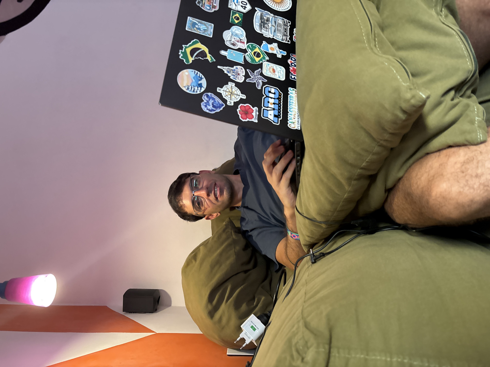 A person is seen using a laptop with various stickers on it. The laptop sits on their lap, and they appear to be seated in a beanbag chair. A charging cable is visible near the person. The wall behind the person is painted in white and orange. A lamp with a pink bulb is also visible in the background, along with a black box. The stickers on the laptop include Brazilian flags and references to locations like Florianópolis. Given the content of the stickers, it is likely that the person is in Brazil or has a connection to the country. Without further contextual clues, it is not possible to confirm that this photo was taken in São Paulo, Brazil.