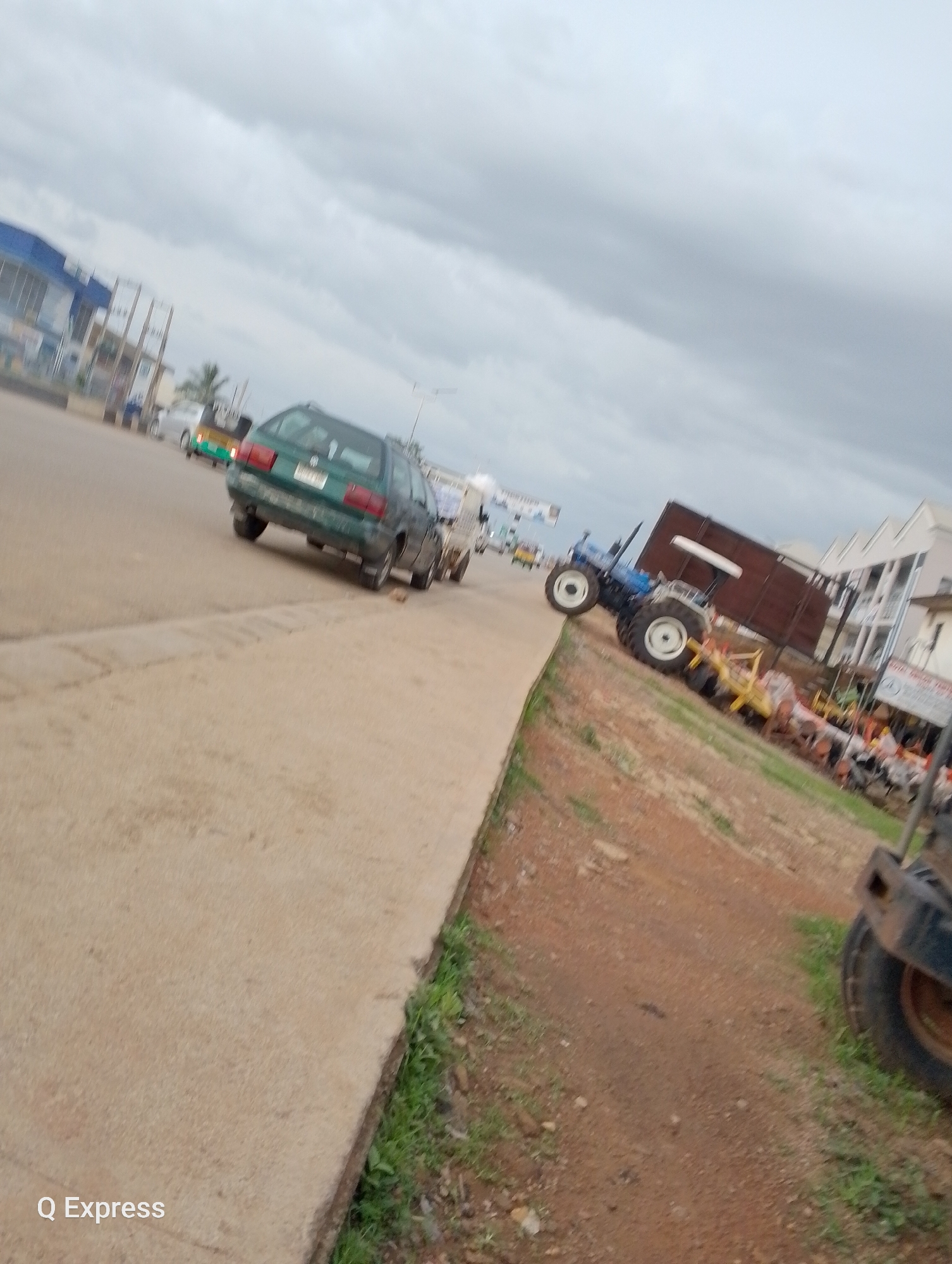 A low-angle, slightly blurry, horizontal shot captures a street scene under an overcast sky. The foreground features a concrete sidewalk with a strip of green grass and red dirt along its edge. A dark-colored, heavy-duty vehicle, possibly a construction machine, is partially visible on the right side of the image, with a large tire dominating the lower right corner.
In the mid-ground, a green station wagon is driving away from the camera, with another small vehicle, possibly a tuk-tuk, behind it. Further down the road, a large blue tractor with a trailer is parked, loaded with a dark brown, rectangular structure. Several agricultural implements are visible near the tractor, suggesting a sale or display of farming equipment.
In the background, buildings line the street, including a blue and white structure on the left that appears to be a commercial establishment. The sky is filled with dense, gray clouds, indicating cloudy or possibly rainy weather. The overall lighting suggests it is daytime, likely morning or afternoon. The image seems to be taken in an urban or semi-urban area in Jos, Nigeria, given the presence of common vehicles and the overall environment. No clear emotional cues are evident due to the blurriness and focus on vehicles and scenery. The text "Q Express" is visible in the bottom left corner of the image, likely a watermark.
