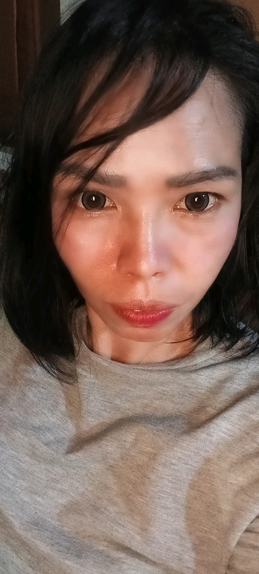 A close-up, frontal portrait shows a person's face and upper torso. The subject has dark hair that falls across their forehead, obscuring part of their face. They are wearing a grey t-shirt. The lighting in the image is warm and casts a glow on the subject's skin, highlighting areas of their cheeks and nose. The background is dark and indistinct, with some subtle textures visible, possibly indicating a curtain or wall. The image appears to have been taken indoors, and there is no information to suggest a location in Kuala Kapuas, Indonesia.
