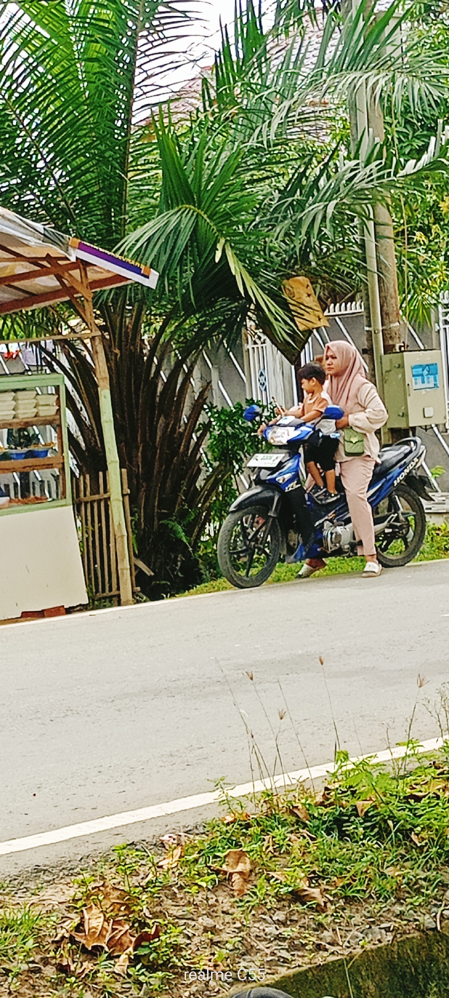 In Lhokseumawe, Indonesia, a woman wearing a light pink hijab and matching outfit is riding a blue motorcycle. She is seated on the motorcycle, which is stopped on the side of an asphalt road. Next to her, in front of her on the motorcycle, is a young boy wearing a light brown shirt and dark pants. He is facing forward, looking ahead. The woman is holding the handlebars of the motorcycle with her right hand and appears to be looking forward as well. She is carrying a small green purse on her left shoulder.
The environment is lush and green, with tall palm trees dominating the background. To the left of the motorcycle, a small roadside stall with a wooden structure and a white exterior is visible. The stall has shelves displaying white containers. The ground beside the road is covered in grass, fallen leaves, and small stones. The sky is overcast, suggesting a possibly humid day. The road has a white line marking its edge. A utility box is mounted on a pole behind the woman. The scene appears to be a typical roadside view in a tropical urban or suburban area.