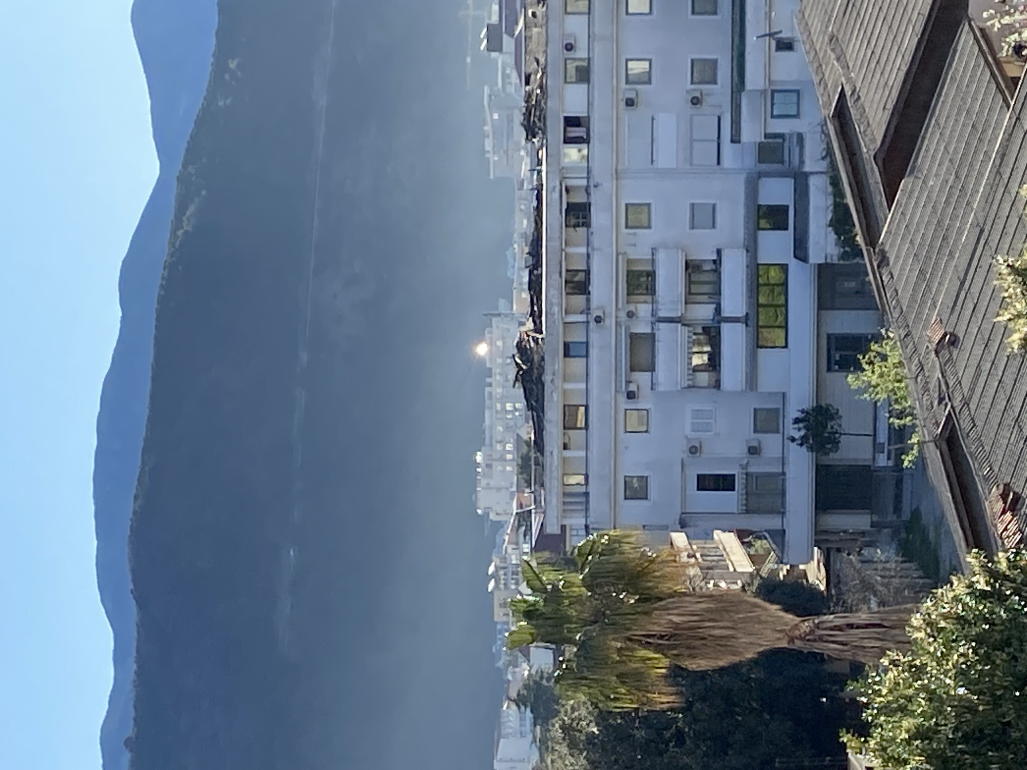 The image shows a daytime scene in Budva, Montenegro, featuring mountains on the left, a cityscape in the center, and part of a building on the right. The mountains are dark and hazy, contrasting with the blue sky. The cityscape includes white buildings with numerous windows, some with visible air conditioning units. A palm tree and other greenery are visible in the foreground, partially obscuring the lower part of the buildings. On the right, a building with a dark, steeply angled roof is present. The sun reflects off a distant building.