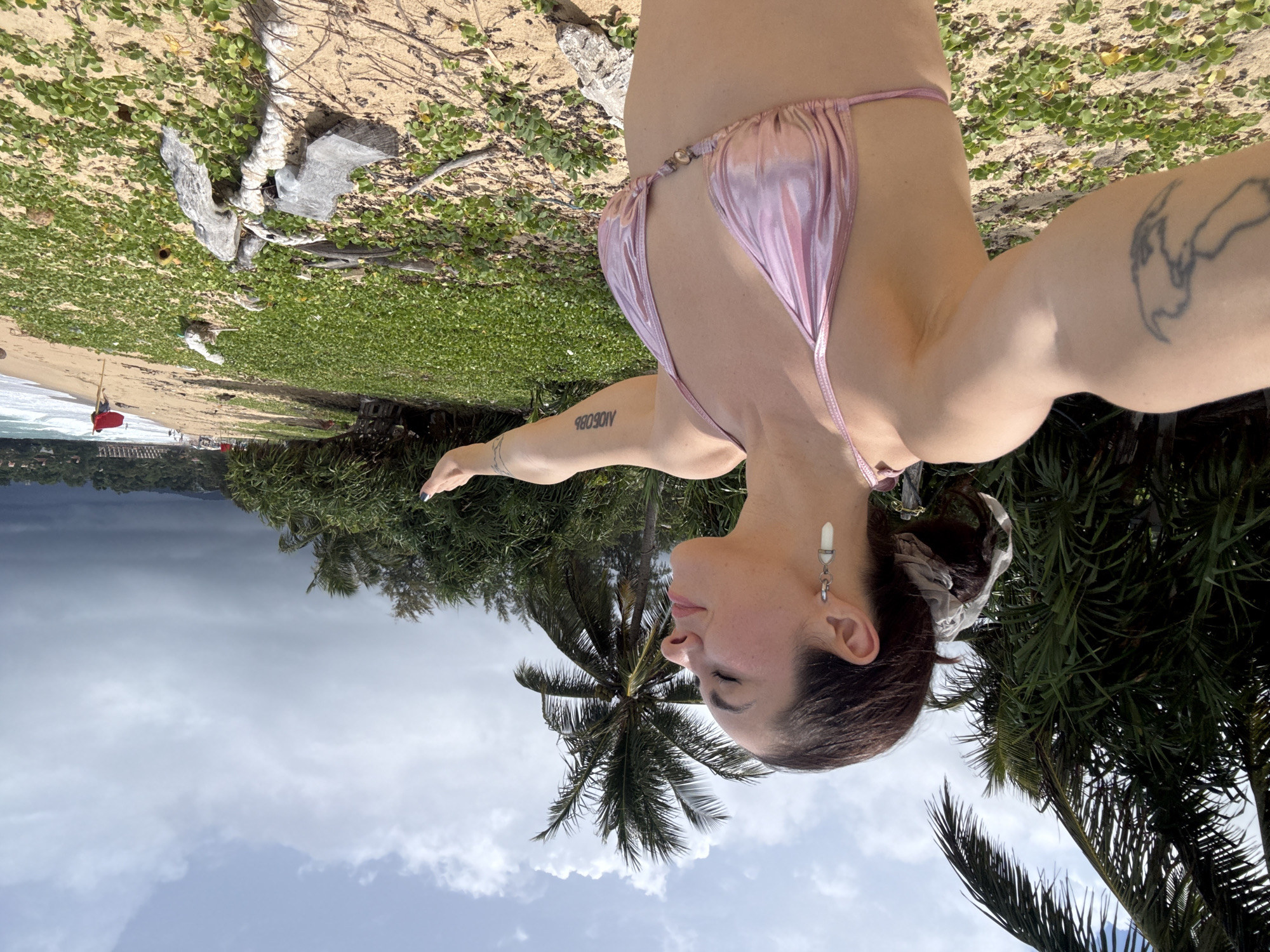 A woman with dark hair pulled back by a scrunchie stands on a sandy beach with her eyes closed and arms outstretched, facing the sea. She is wearing a metallic pink bikini. A tattoo is visible on her left arm. The background features lush green vegetation, palm trees, and the ocean with waves. In the distance, there are other people on the beach and a red flag on a pole. The sky is partly cloudy.