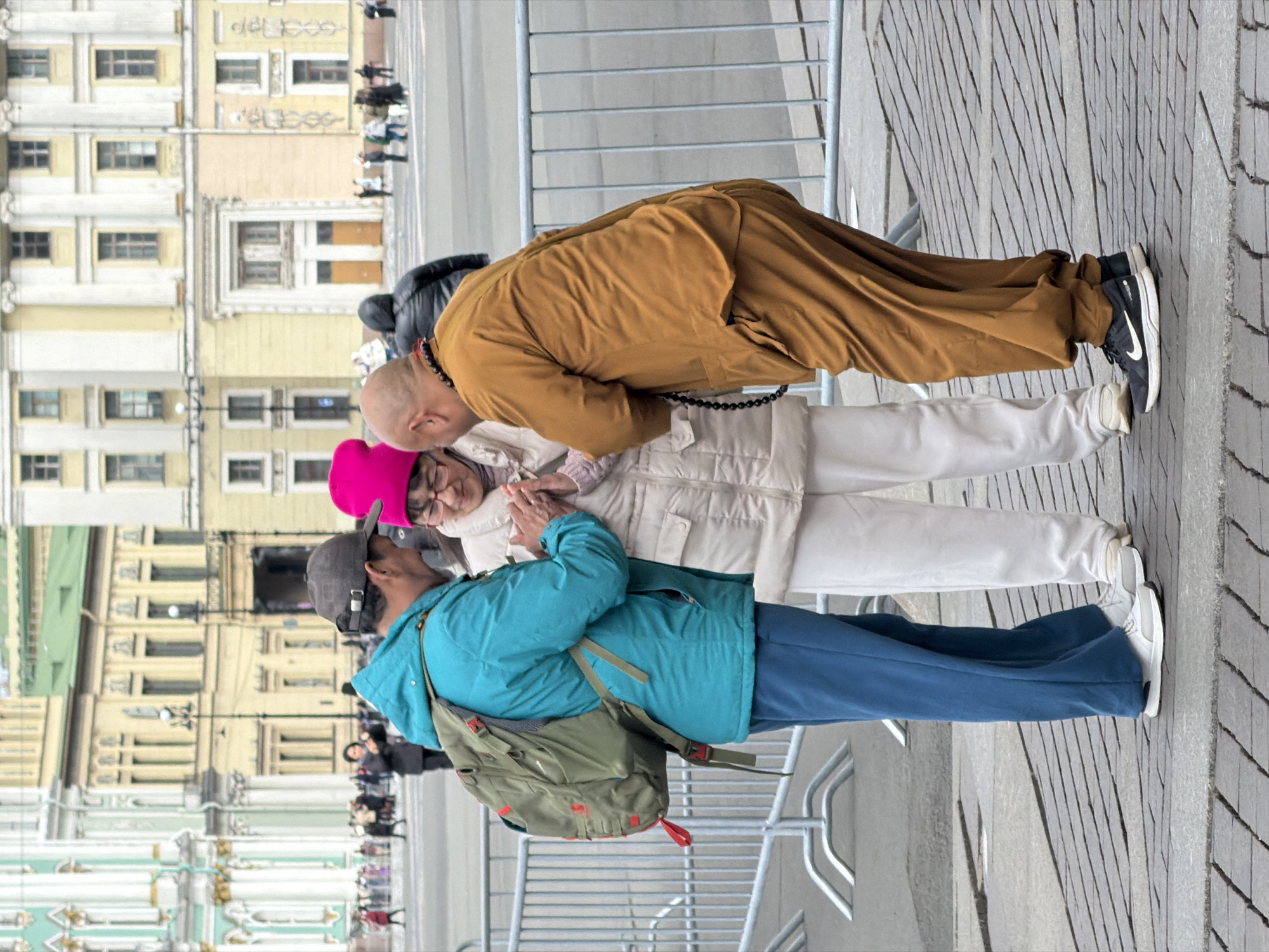 This image captures a street scene in Saint Petersburg, Russia, focusing on an interaction between three individuals in the foreground. The photograph is oriented sideways, but depicts a daytime setting with cool weather.
At the center, a person with a shaved head, possibly a Buddhist monk or practitioner, is dressed in a flowing, earthy-toned (brown/orange) robe with prayer beads around their neck. They are bowing or leaning forward towards an older woman standing beside them. The woman is wearing a light-colored winter jacket and a vibrant pink hat, with glasses on her face. Her hands are clasped together in front of her, a gesture often associated with prayer or reverence, as she looks towards the robed individual. Adjacent to them, slightly behind, stands another person in a teal blue jacket, blue pants, and a grey baseball cap, carrying a green backpack. This individual appears to be observing the interaction.
The background features stately, multi-storied buildings with numerous windows and classical architectural details, predominantly in light yellow and green tones, characteristic of Saint Petersburg. Metal barriers or fences run horizontally across the middle ground, separating the main subjects from other blurred figures in the distance, indicating a public space or thoroughfare. The ground is paved with patterned grey stones. The attire of the individuals suggests cool weather, and the soft lighting indicates it is daytime, possibly overcast. No discernible text is visible.