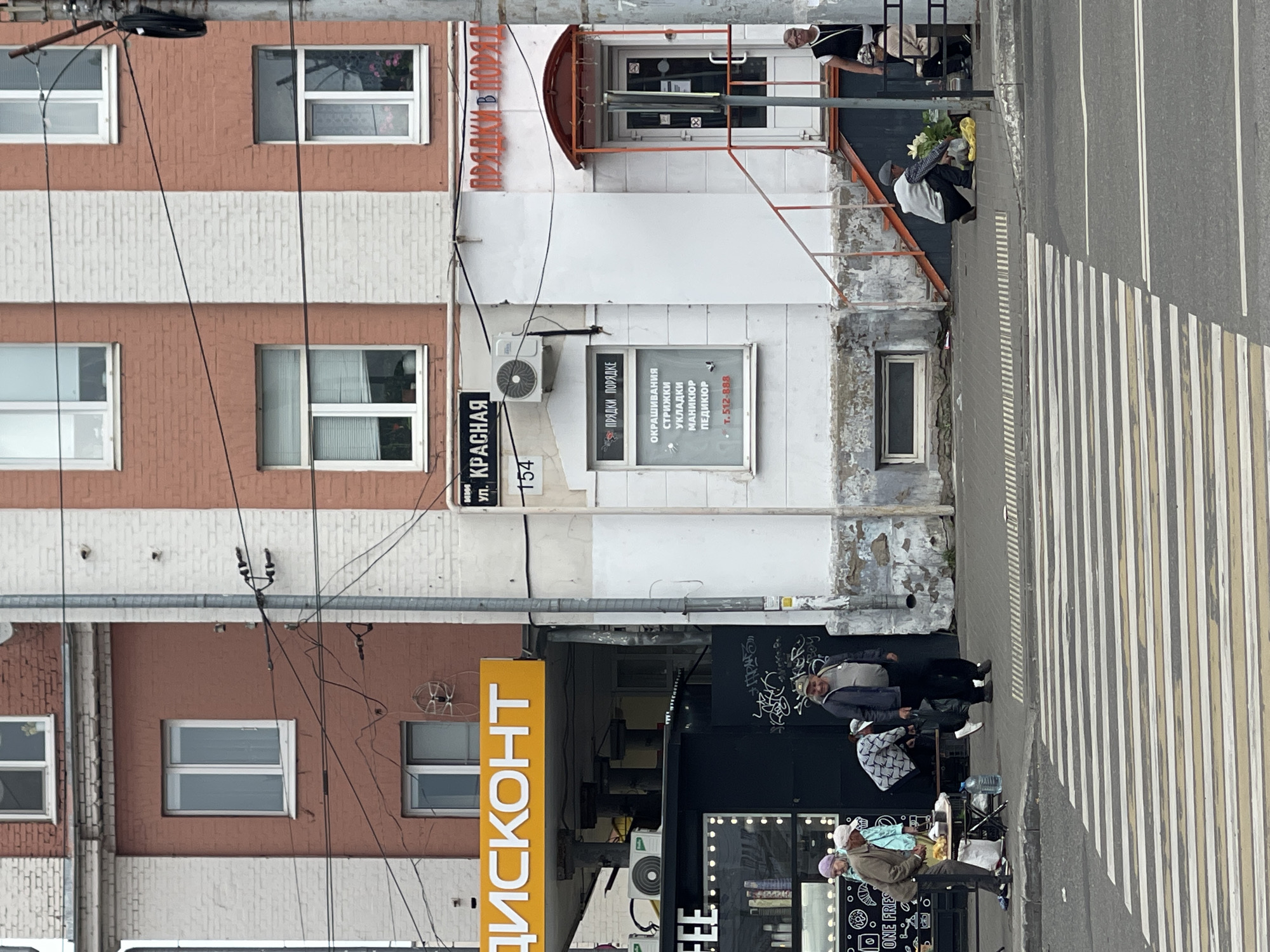 The image depicts a street scene in Izhevsk, Russia, with a focus on a section of a multi-story brick building. The building features red and white brickwork, with several windows visible. One prominent sign reads "ДИСКОНТ" (Discount) in yellow lettering against a yellow background. Below this sign, a black kiosk with the word "FFEE" is partially visible, with some items displayed inside.
On the sidewalk in front of the building, there are several people. Three individuals, two men and a woman, are gathered around a table laden with items, possibly food or goods for sale. A fourth person, a man in a blue jacket, is walking away from the table towards the right of the frame. Further to the right, a man in a grey cap is crouching down near a pile of flowers on the sidewalk. Another woman, wearing glasses and a black top, stands on the sidewalk to the right, observing the scene.
A crosswalk with yellow and white stripes is prominent in the foreground, indicating a pedestrian crossing. The environment appears to be an urban setting with visible power lines and a streetlamp. There is graffiti on the lower part of the building's facade. A sign on the building reads "УЛ. КРАСНАЯ" (Krasnaya Street) and "154". Another sign above a doorway indicates services like "ОКРАШИВАНИЕ" (coloring), "СТРИЖКИ" (haircuts), "УКЛАДКИ" (styling), "МАНИКЮР" (manicure), and "ПЕДИКЮР" (pedicure), with a phone number "Т. 512-888". There is also a sign that says "ПРЯДКИ В ПОРЯДКЕ" (Hairdressing in order) in red lettering.