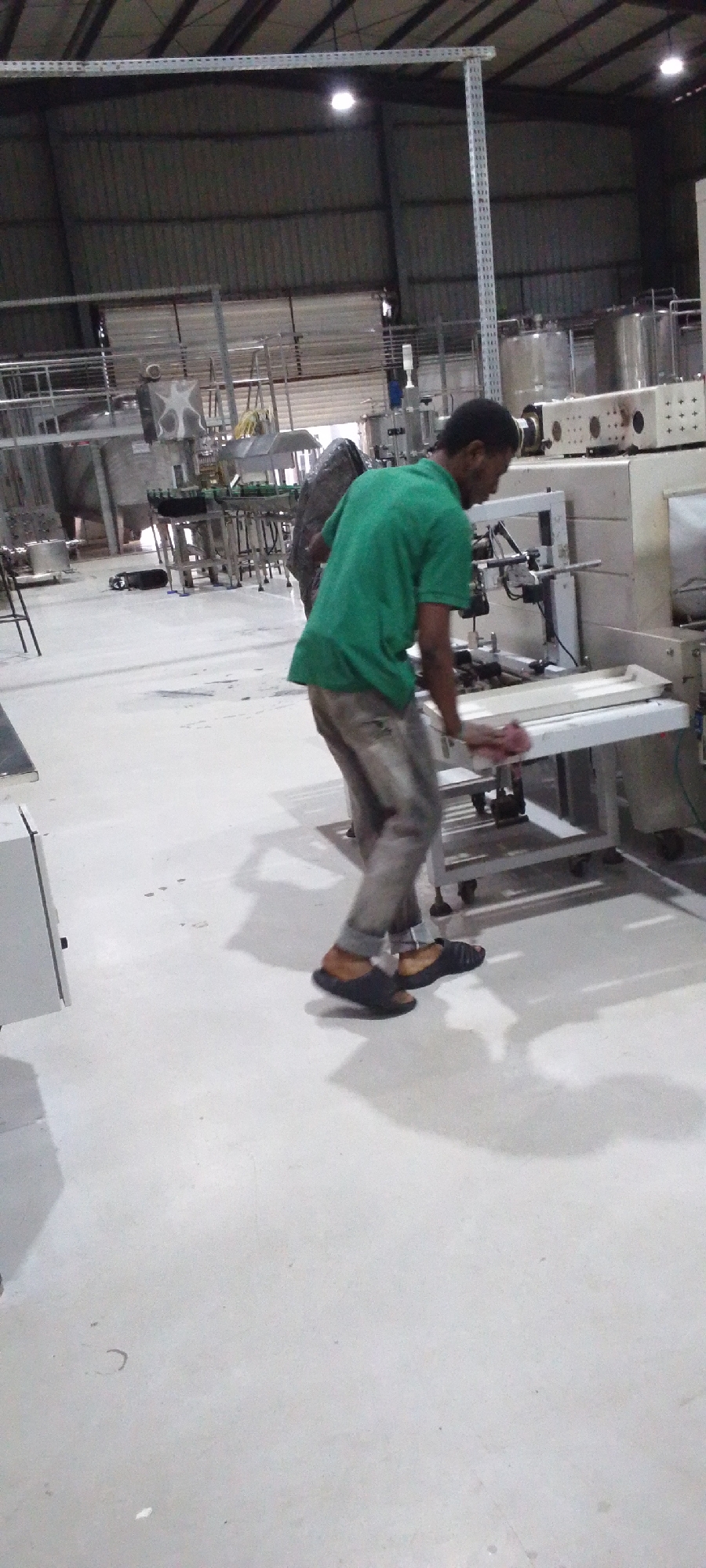 The image shows a man in a green t-shirt and grey pants standing in a large, industrial-looking space. He is cleaning a white table-like structure with a pink cloth. The area appears to be a manufacturing or processing facility, with various pieces of equipment and machinery visible in the background. He is wearing open sandals. There are liquid storage tanks. There is one other man present. The location of the photo is Unknown, Unknown.