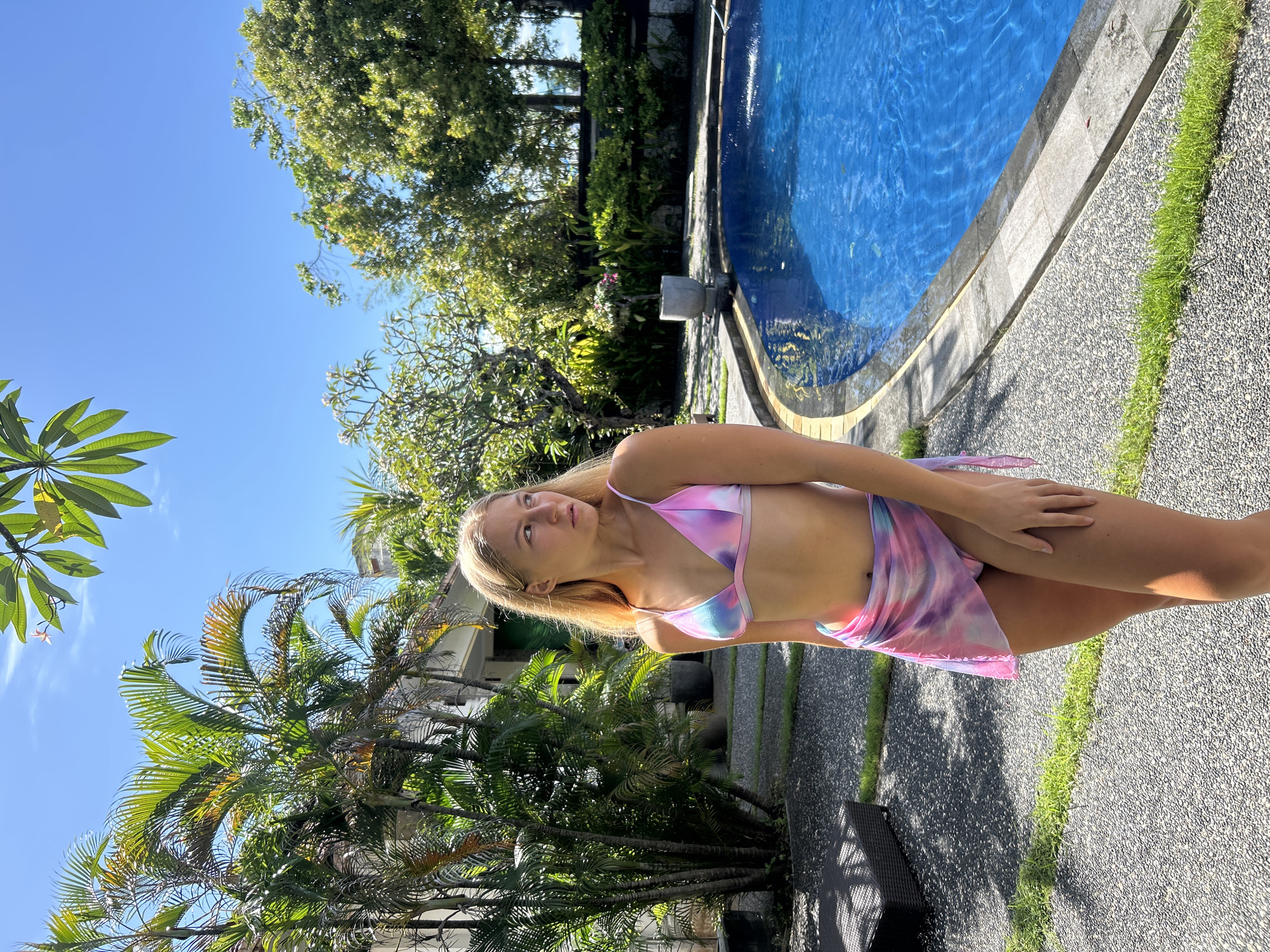 A full-length shot shows a blonde woman in a pink and blue tie-dye bikini and wrap, standing by a swimming pool on a sunny day. The location appears to be outdoors in a tropical environment, characterized by palm trees and lush greenery, possibly in Kuta Selatan, Indonesia, due to the environment. The pool's edge and surrounding pathway suggest a resort or private residence setting. The woman is looking towards the sky.