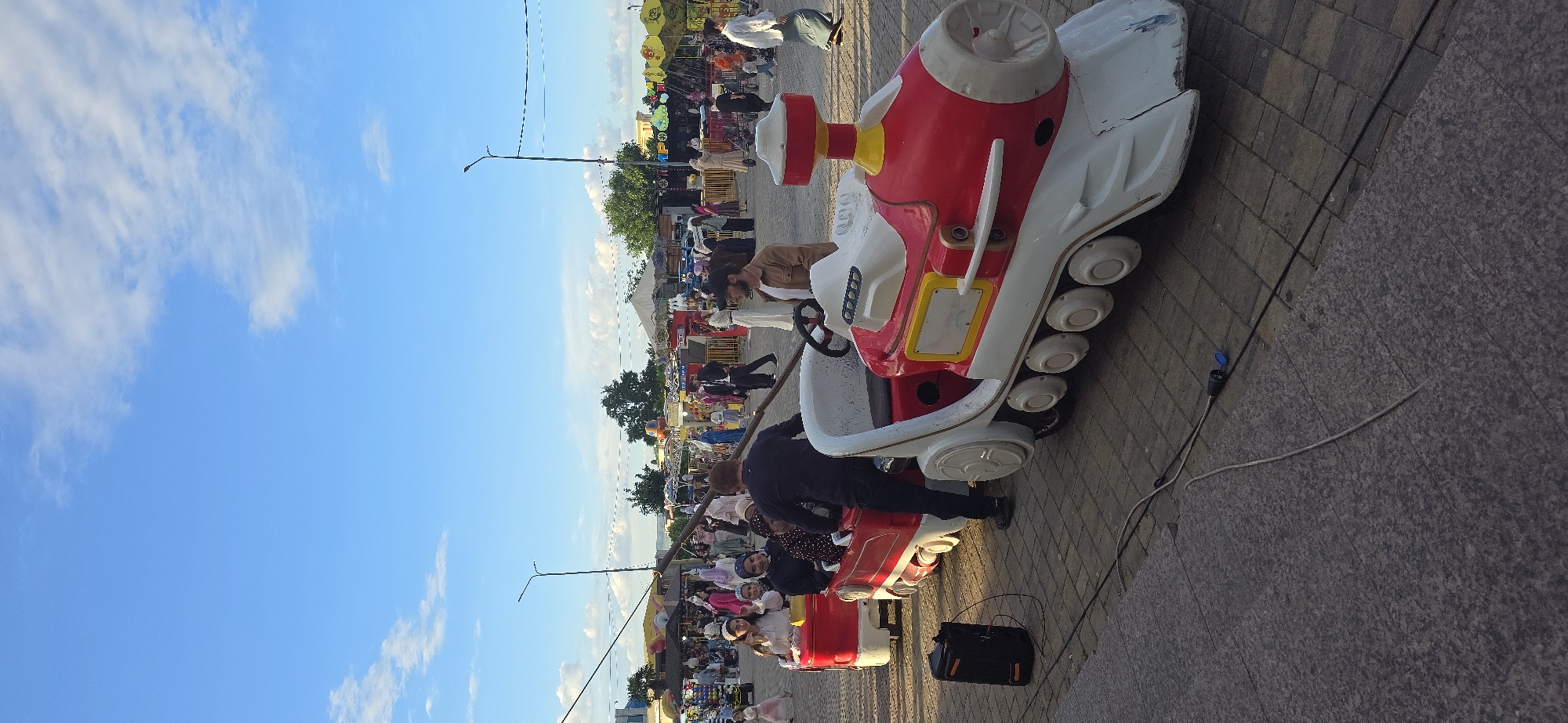 The image depicts an outdoor scene in what appears to be a park or public space, likely in Grozny, Russia, based on the style of architecture and public amenities present. The sky is blue with scattered clouds. Several rides and stalls are set up, indicating a fair or market. Numerous people, including adults and children, are visible, with some riding on coin-operated vehicles such as a train ride. One man is tending to the ride, while a bag sits close by. The ground is paved with stone or tile.