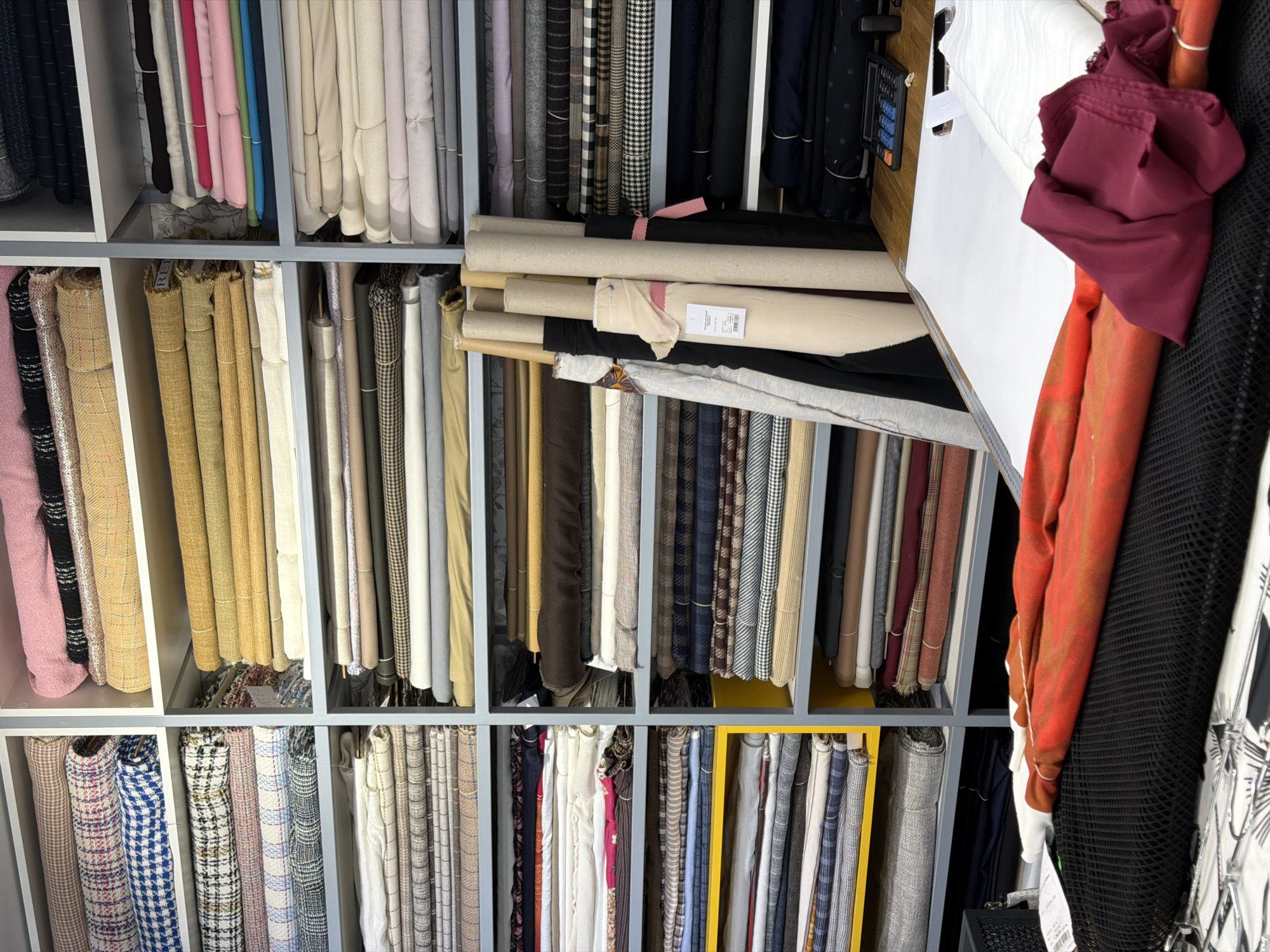 The image displays the interior of a textile store or warehouse, specifically located at Efimova street, building 3, in Saint Petersburg, Russia, as evidenced by a label on a fabric roll. The scene is dominated by numerous floor-to-ceiling shelving units, predominantly white and light grey, with some yellow panels on the lower left section. These shelves are meticulously filled with a vast array of fabrics, neatly organized into stacks and rolls.
Fabrics exhibit a wide range of colors, patterns, and textures, including various solid colors such as pink, blue, green, beige, brown, grey, black, and white, as well as patterned textiles like plaids, houndstooth, and stripes. In the mid-ground, several large fabric rolls, some wound around cardboard cores, stand vertically, leaning against a shelving unit.
The foreground features a wide, white work surface or cutting table. On this table, a selection of fabrics is laid out or draped, including a piece of black mesh-like fabric, a mottled orange and red fabric, and a folded burgundy-colored fabric. A small white fabric bundle is also present. To the right on the table, a black calculator and another small dark electronic device are visible. No individuals are present in the image.