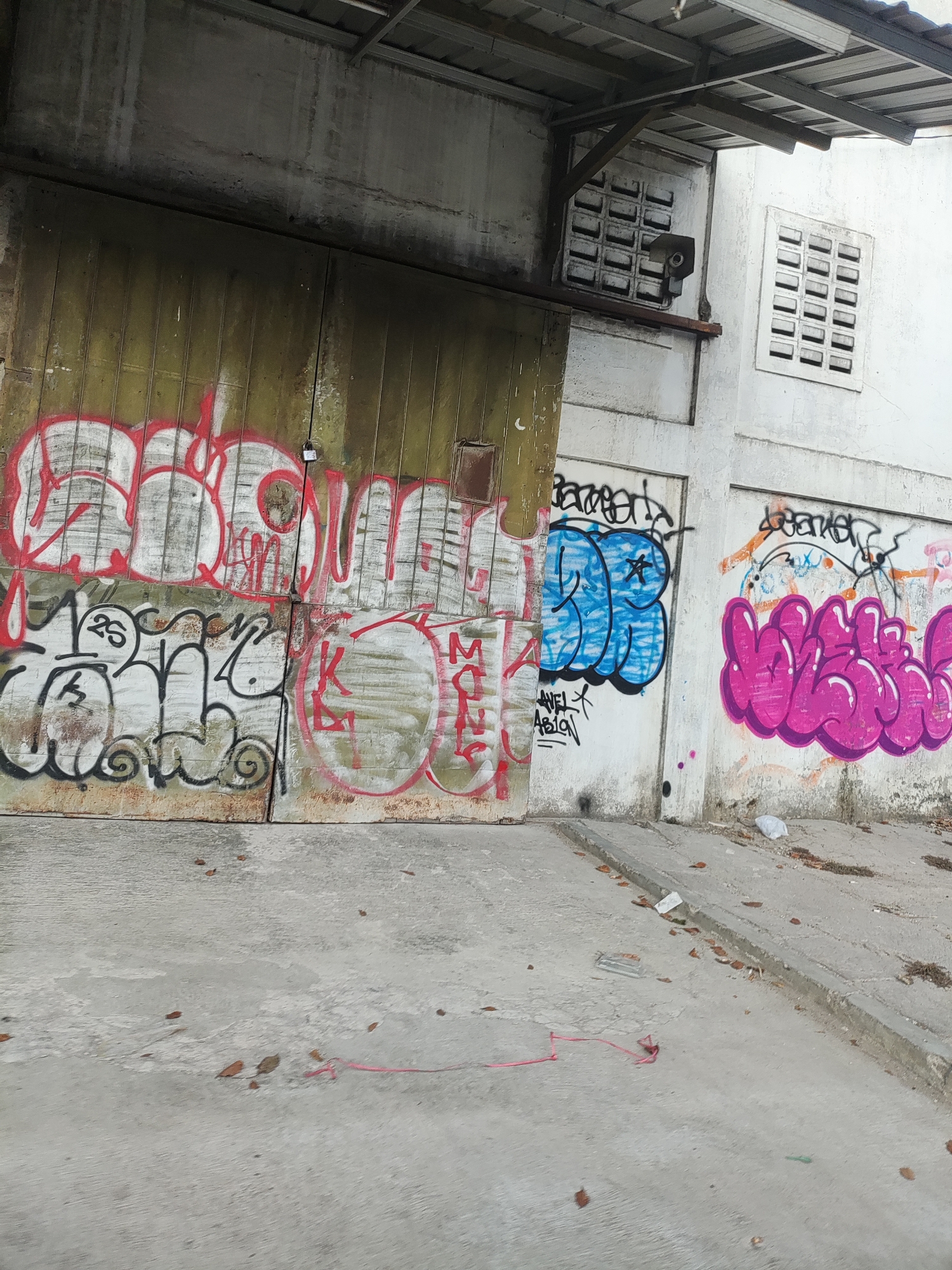 The image depicts a section of an outdoor urban environment in Cirebon, Indonesia. The scene is characterized by a large, weathered metal garage door covered in red and white graffiti, with additional graffiti in black and white below it. To the right of the garage door is a white wall with more graffiti, including blue bubble letters and pink bubble letters, as well as some smaller tags. There are two louvered vents visible on the white wall, one of which has a small camera mounted to it. The ground in front of the wall and door is concrete and appears somewhat worn and dirty, with scattered leaves and debris. A thin pink cord or wire is visible on the concrete surface in the foreground. The overall impression is one of urban decay and street art.