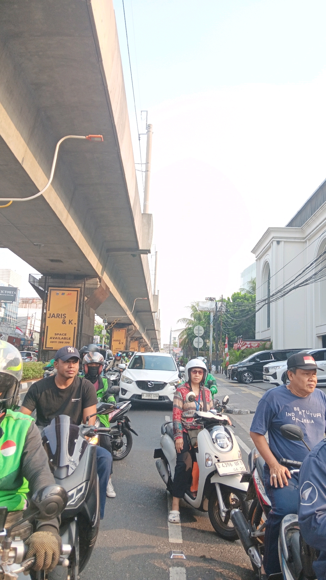 The image captures a traffic scene in South Jakarta, Indonesia, under an elevated highway structure. In the foreground, several individuals are seated on motorcycles, engaged in traffic. A man in a black t-shirt and a baseball cap is positioned on the left, looking forward. To his right, another person wearing a helmet and a green vest is also on a motorcycle. Further to the right, a woman with a white helmet and sunglasses rides a white scooter. She is wearing a patterned long-sleeved shirt and black pants. To the far right, a man in a blue t-shirt is partially visible, also on a motorcycle.
Several vehicles are present on the road, including a white Mazda CX-5 SUV in the middle ground. The road is paved with lane markings and reflective road studs. Overhead, the concrete structure of an elevated highway dominates the upper portion of the image, with visible signage attached to its support pillars. One sign, in yellow, displays the text "JARIS & K." and "SPACE AVAILABLE" with a phone number. In the background, buildings and lush green trees are visible, typical of an urban environment. The sky is bright and appears to be clear. The overall atmosphere suggests a typical day with moderate traffic flow.