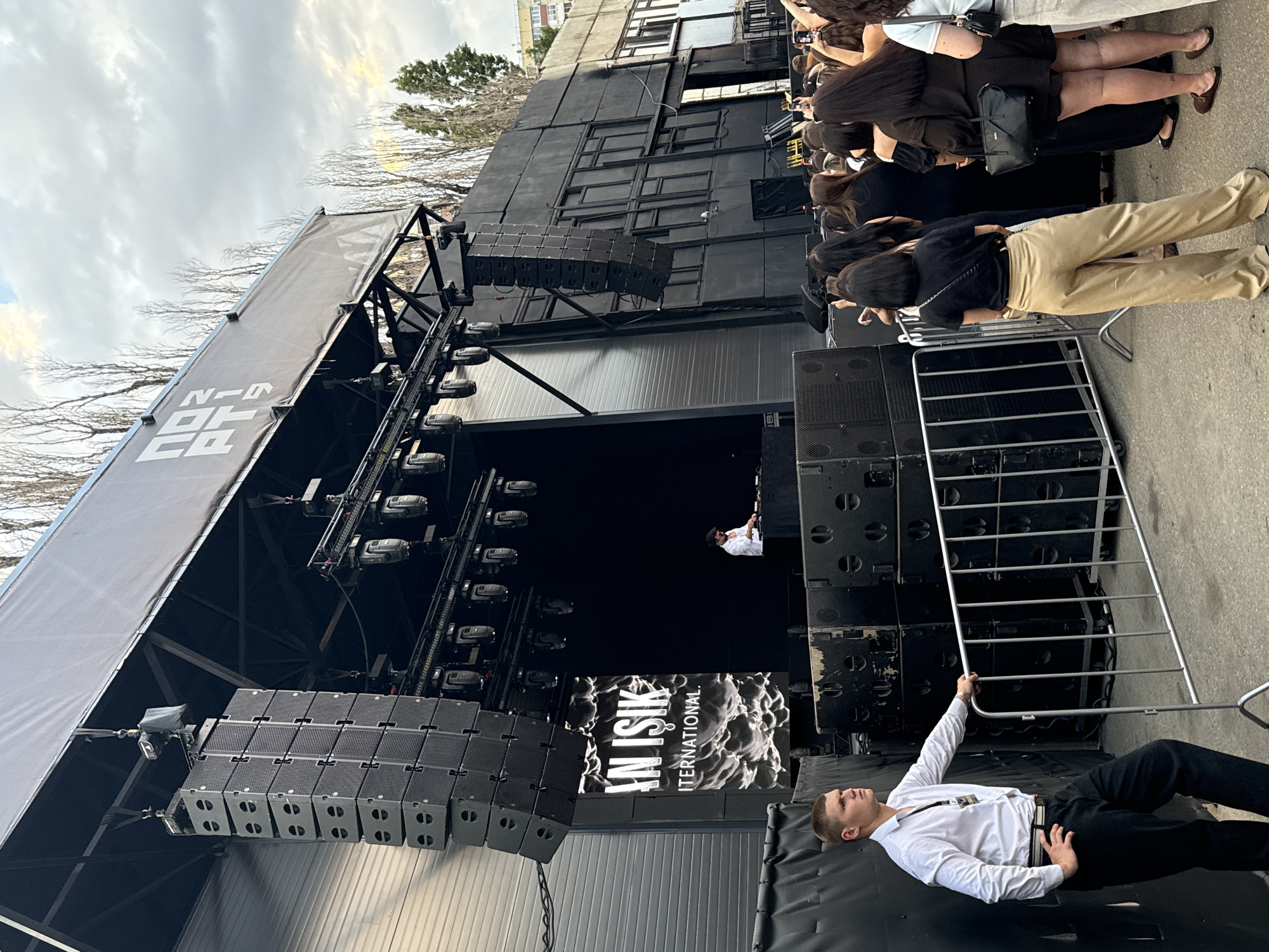 The image depicts an outdoor event stage, likely in the city of Kozelsk, Russia, given the context. In the foreground, a security guard in a white shirt and black pants stands near a metal barrier, looking to the left. Behind him, a crowd of people, mostly women, are gathered, facing the stage. The stage itself is large and black, with rows of speakers stacked on either side. A banner displaying "AN ISIK INTERNATIONAL" hangs prominently on the left side of the stage. A DJ is visible on the stage, operating equipment. Above the stage, a large black structure with the logo "NO2 PT.9" is mounted. The background shows bare trees and a cloudy sky, suggesting an autumn or early spring setting. The overall scene suggests an outdoor concert or festival.