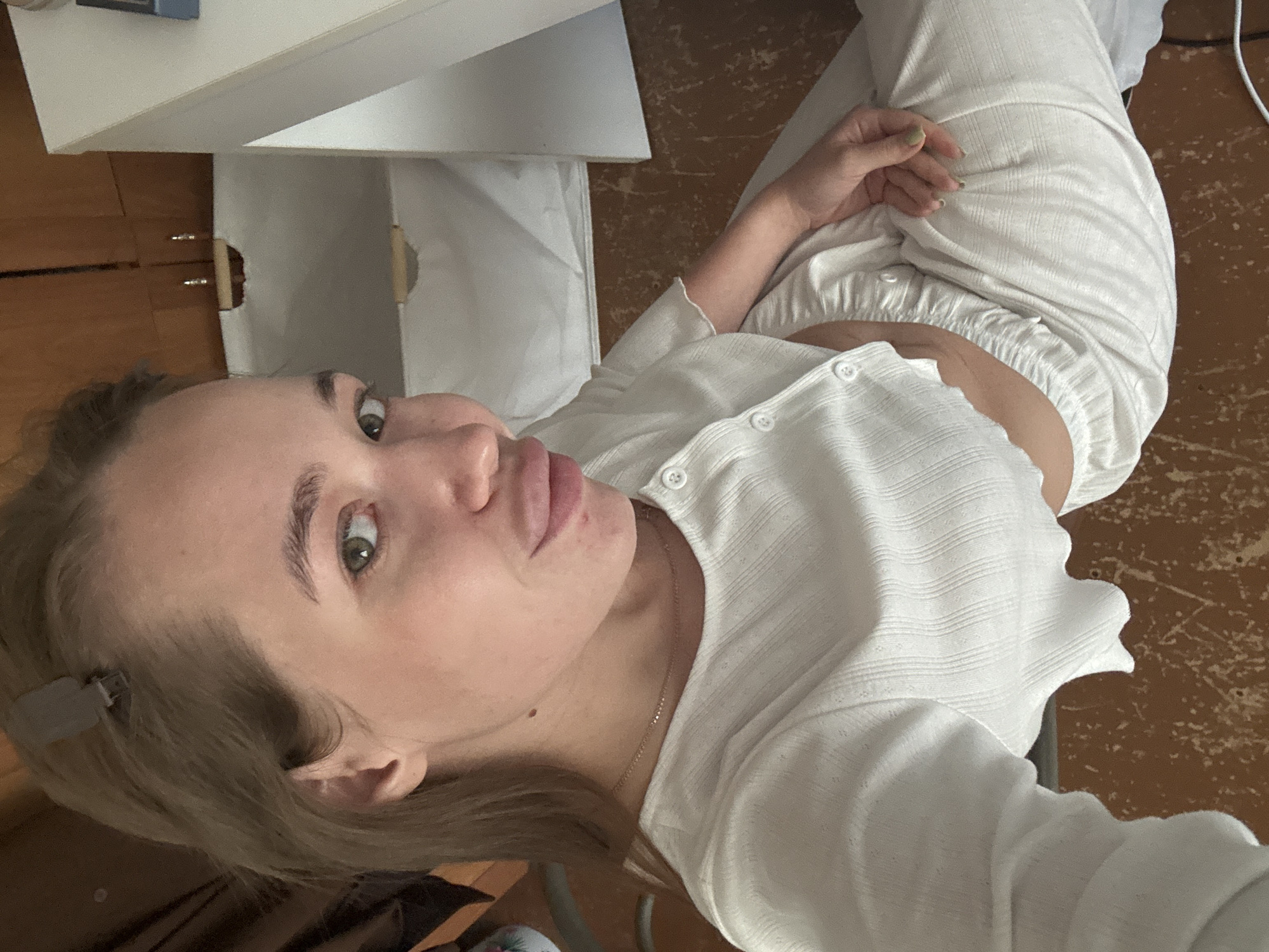 A young woman with light brown hair, pulled back with a clip, is sitting indoors and taking a selfie. She is wearing a white ribbed top and matching pants. Her gaze is directed towards the camera, and her lips are slightly parted. The background shows wooden cabinetry and a white surface, possibly a desk or shelf, with some laundry or fabric items visible within an open compartment. The floor appears to be linoleum or wood with visible wear. The lighting suggests an indoor setting, possibly a home. The image does not provide any specific indicators of the location being Chaikovsky, Russia.