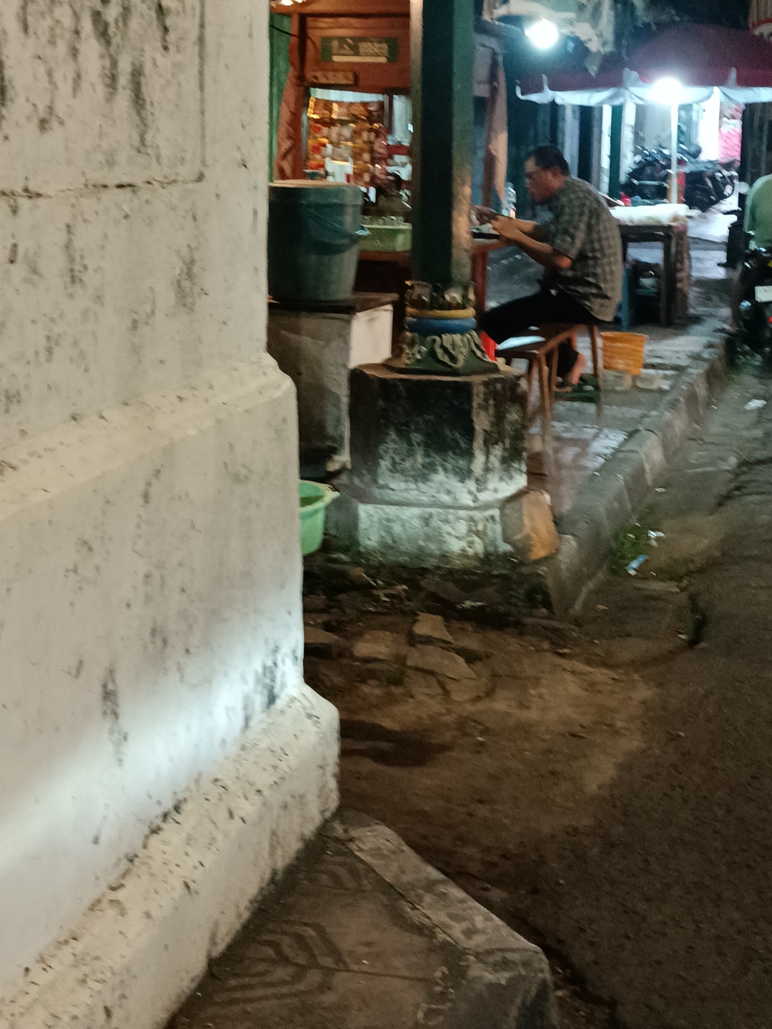 A man in a checkered shirt sits on a stool at an outdoor food stall in Yogyakarta, Indonesia, appearing to eat or prepare food. He is facing a table, and is illuminated by overhead lights. Behind him, motorcycles are parked and several umbrellas are open, suggesting it is nighttime or the stall is covered. A large white pillar is in the foreground, and a green bucket sits on a stone block to the man's left. The ground is paved with rough stones, and there is some debris on the ground.