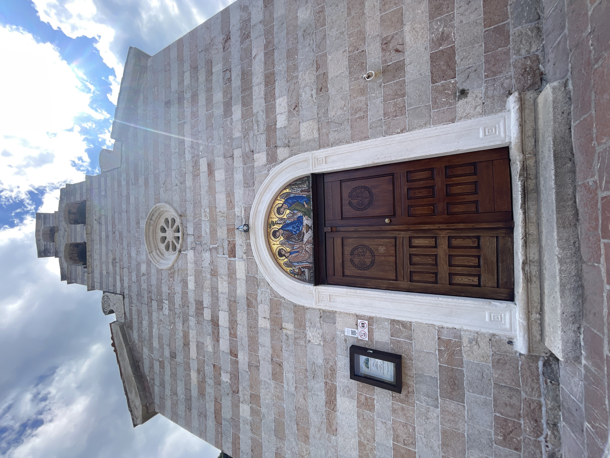 The image captures the exterior of a stone building, seemingly a church, in an unidentified location. The facade is constructed of striped gray and brown stone bricks, creating a textured surface. The main entrance features a wooden door adorned with intricate patterns, set within a white stone frame. Above the door, a semi-circular mosaic panel adds a colorful detail. Adjacent to the entrance, a framed display with printed information is mounted on the wall. A circular window is located higher up on the building's facade. The architectural style suggests a European, possibly Italian, setting. The cloudy sky is visible in the background.