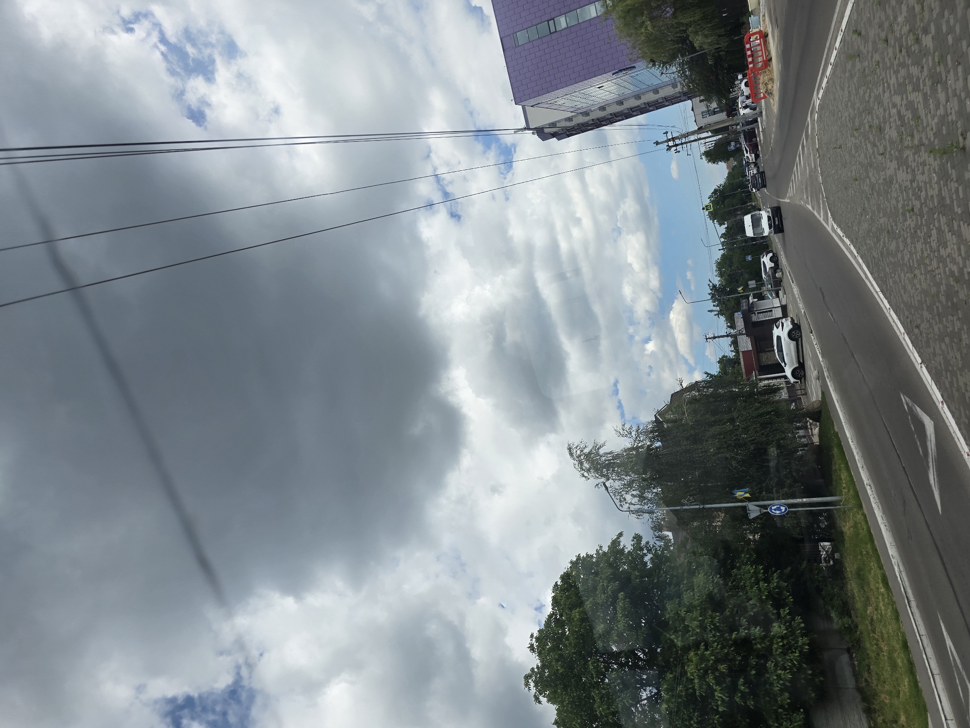 The image captures a street scene in Petropavlivska Borshchahivka, Ukraine, on a cloudy day. A paved road with white lane markings curves through the scene. Several vehicles, including a white van, are visible on the road, indicating moderate traffic. On the left side of the road, there are trees and buildings, some with grass in front. On the right side, a modern purple-tiled building stands out, with more trees and construction barriers nearby. Power lines stretch across the sky, which is dominated by large, gray clouds interspersed with patches of blue sky and white clouds. The overall impression is a typical urban or suburban environment under a changeable sky.