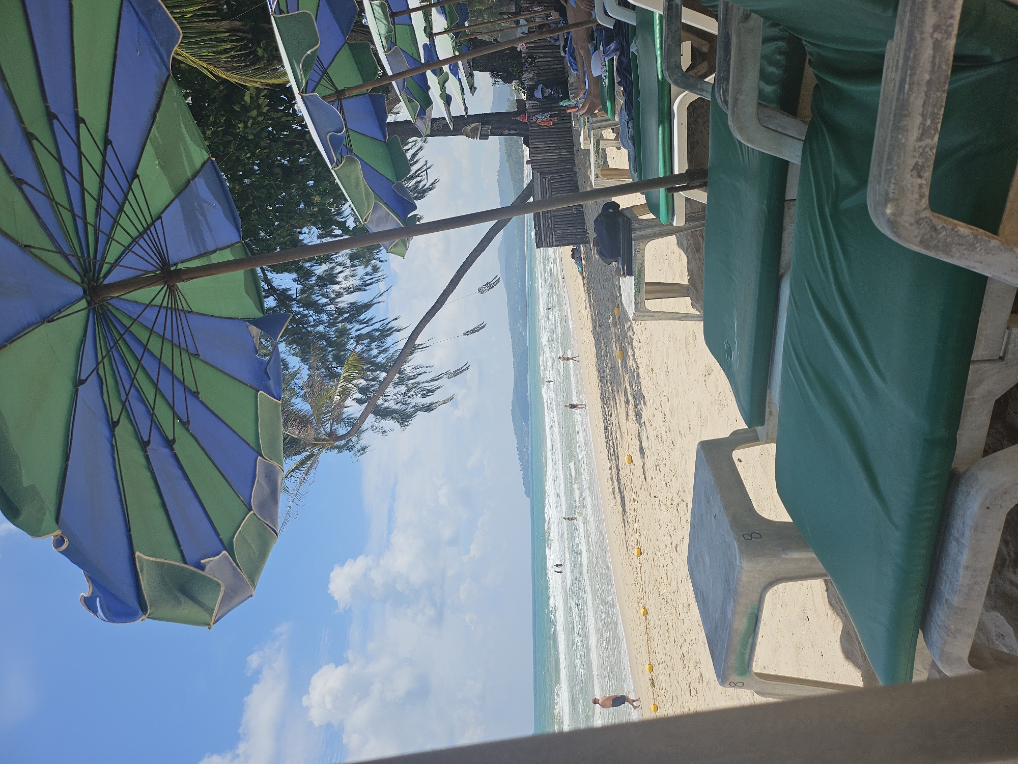 The image captures a tropical beach scene in Choeng Thale, Thailand, under a bright, partially cloudy sky. In the foreground, several green-covered lounge chairs are arranged on the sandy beach, some with white plastic frames. A small, square, light-colored plastic table with the number "8" is positioned in front of one of the chairs. A large, blue and green striped beach umbrella is prominently displayed, angled to provide shade. In the middle ground, more lounge chairs and umbrellas are visible, with a low wooden fence bordering the area behind them. A few people are scattered across the beach and in the shallow, turquoise ocean, with gentle waves breaking on the shore. In the background, a distant coastline with hills is visible. The overall environment suggests a relaxed atmosphere at a seaside resort.