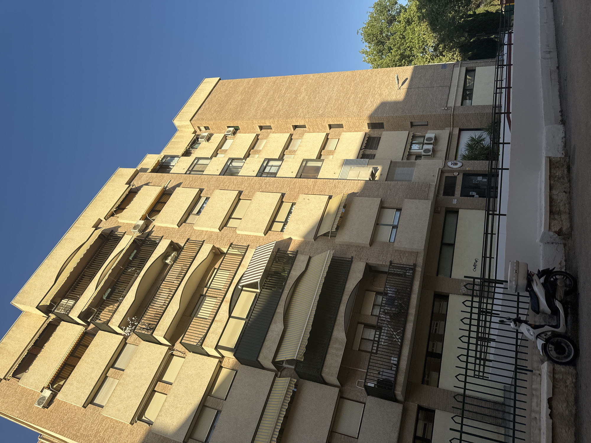 The image shows a tall, multi-story brick building with balconies and awnings, set against a clear blue sky. A white scooter with a storage box is parked in front of a black metal fence. The building appears to be an apartment complex, with several windows and air conditioning units visible. The brickwork is a warm, light brown color, and the concrete balconies are textured. The awnings on the balconies are various shades of yellow and white, with some striped. The scooter is parked on a paved surface, and behind the fence, there is a lighter colored wall with some graffiti. A tree with green foliage is visible to the right of the building. The scene suggests an urban residential area bathed in sunlight.