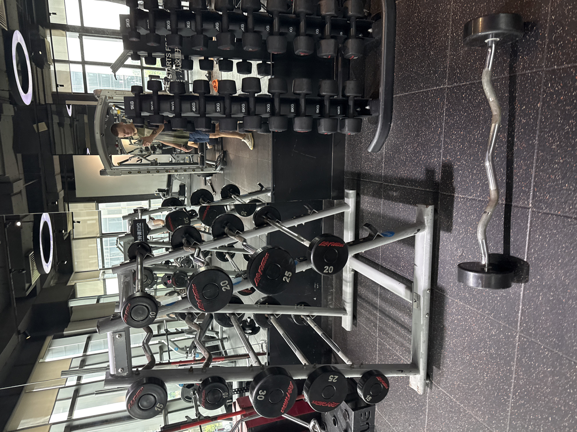The image depicts the interior of a modern gym located in Haizhu District, China. A young man, wearing a dark green t-shirt and blue shorts, stands in front of a large mirror, holding a smartphone and seemingly taking a selfie. He is positioned between a rack of dumbbells and weight plates.
The gym features numerous weightlifting implements. To the left, a rack holds a variety of weight plates, some of which are labeled with weights like "10," "15," "20," "25," and "30." These plates are black with red and white lettering. Behind this rack, another set of weight plates is visible.
To the right of the man, a rack displays a range of dumbbells, neatly organized by weight. The dumbbells are black and have labels such as "1KG," "2KG," "3KG," "4KG," "5KG," "6KG," "7KG," "8KG," "9KG," and "10KG." A sign on the dumbbell rack has text in both English and Chinese, with the English portion reading "CE STARTS FR..." and below it "...从这里开始."
In the foreground, a curved barbell with black weights on each end lies on the dark speckled gym floor. The gym environment is well-lit, with large windows in the background offering a view of buildings, suggesting an urban setting. The ceiling has modern lighting fixtures, including a circular ring light. The floor is covered with a durable, dark rubber matting typical of gym facilities. The overall impression is one of a clean and well-equipped fitness center.