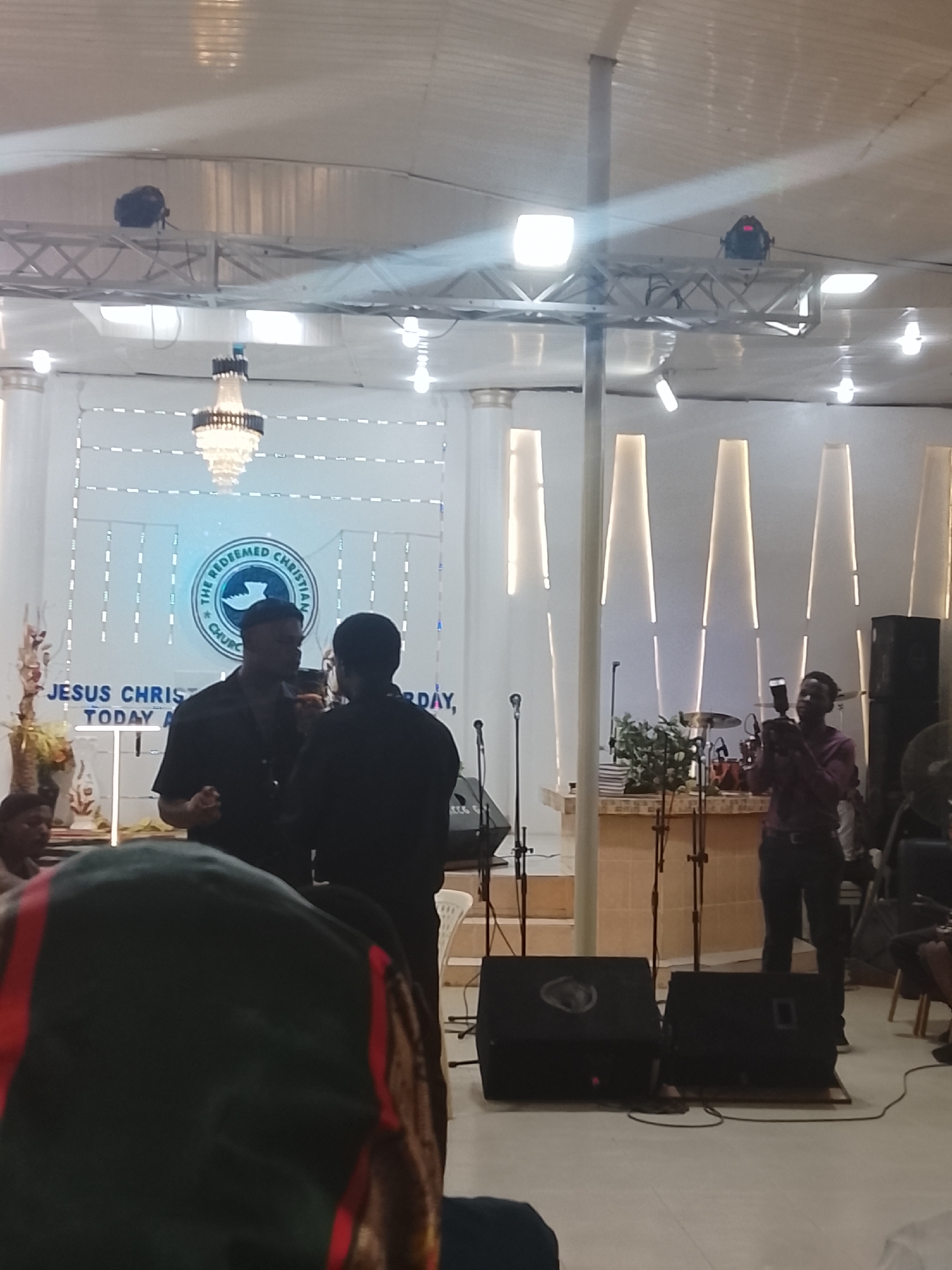The image shows the interior of The Redeemed Christian Church of God, likely in Jos, Nigeria. Two men in dark clothing stand in the foreground facing each other, apparently in conversation. A third man is to the right of the frame operating a camera, possibly filming or taking photos of the event taking place on stage. Audio equipment, including speakers and microphones, are positioned in the foreground. Behind the men is the stage, which features a drum set and other musical instruments. The church logo is prominently displayed on the back wall, above the stage. The interior architecture features modern lighting elements and columns. Several other people are visible in the periphery. The environment seems to be indoors, with artificial lighting in use.