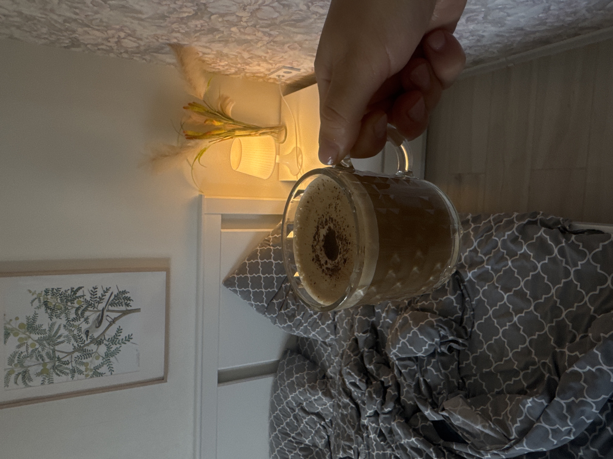 A hand holds a clear glass mug containing a light brown liquid with a dark brown circular design on the foam, over a gray and white patterned quilt on a bed with a white headboard. A framed print of a bird and plant life hangs on the wall to the left of the headboard. A lamp with dried flower arrangements sits atop a white nightstand. The ceiling is white with a textured pattern. The image is rotated counter-clockwise. There is no indication of the location being in Novorossiysk, Russia.