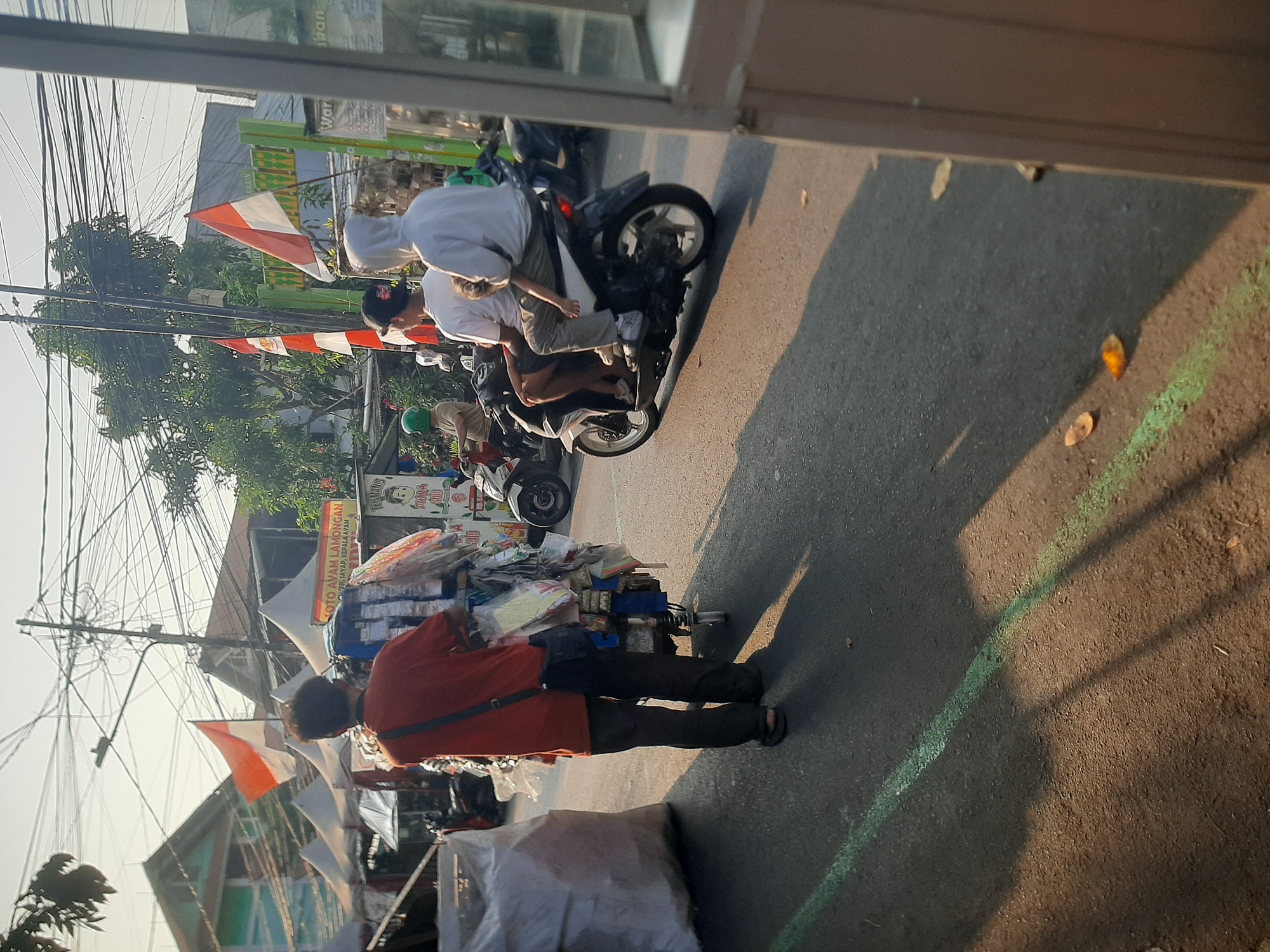 In Central Jakarta, Indonesia, a street scene unfolds under a bright sky with numerous overhead wires. In the foreground, a person wearing an orange shirt and dark pants walks away from the camera, pushing a cart laden with various items, including what appear to be packaged goods. The cart has small wheels and is connected to the person by a strap worn over the shoulder. To the right, a white motorcycle carries three people: a driver wearing a white hoodie, a passenger behind them, and a child seated between them. The motorcycle is in motion, casting a shadow on the asphalt road, which is marked with a faint green line. In the background, several market stalls with white canopies are visible, decorated with Indonesian flags. A sign with text in Indonesian reads "KOTO AYAM LAMONGAN". Other motorcycles are parked near the stalls, and trees are present in the distance. The overall atmosphere suggests a bustling market environment on a sunny day.