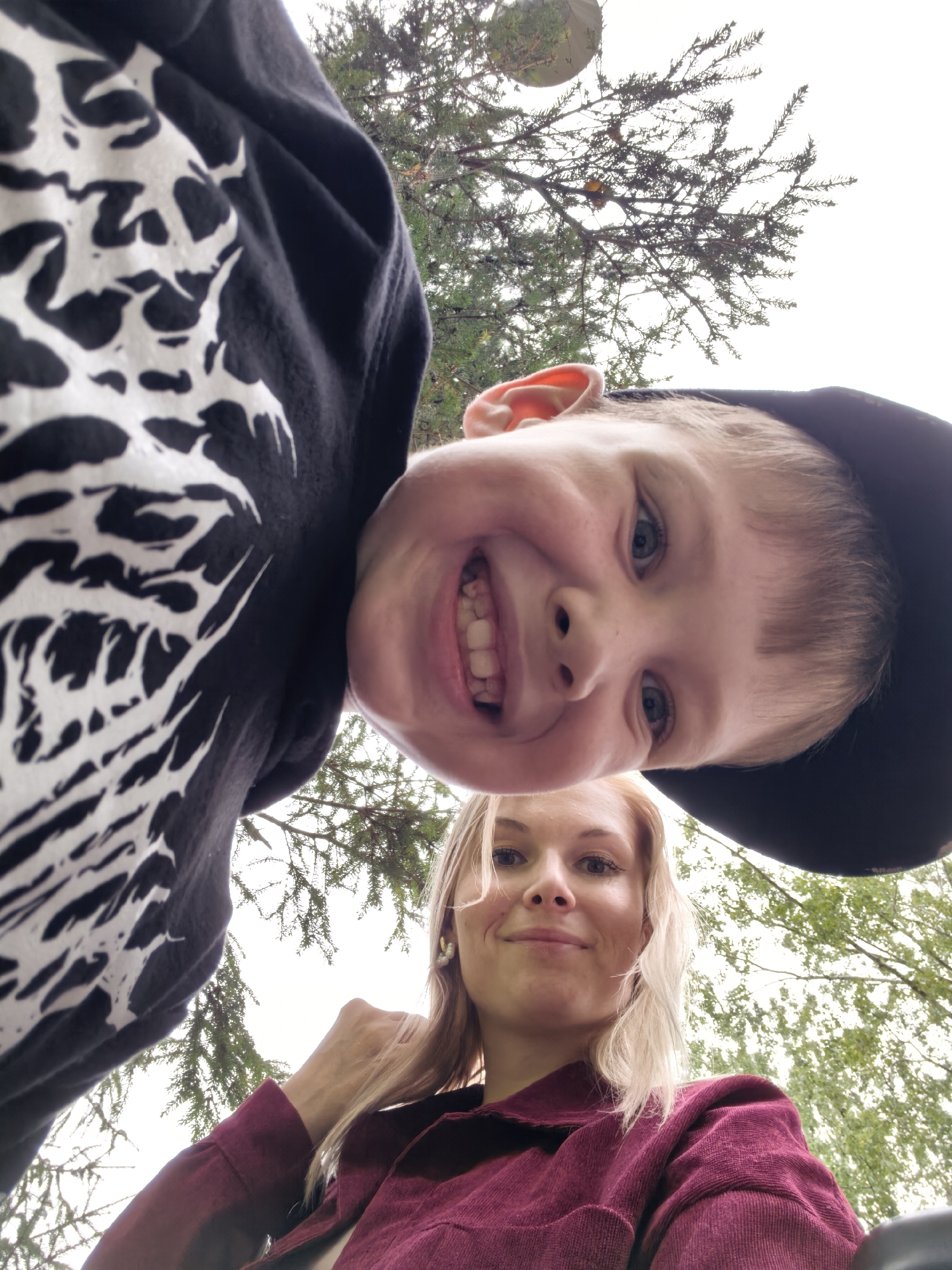 This is a low-angle, upward-facing selfie taken outdoors, likely in Sterlitamak, Russia, under a cloudy sky and amidst the branches of a coniferous tree. Two individuals are visible. In the foreground, a young boy with blonde hair, blue eyes, and a wide smile is looking directly at the camera, wearing a black t-shirt with a white graphic and a black baseball cap. Partially behind and below him, a young woman with blonde hair and light-colored eyes is also looking towards the camera, her right hand touching her hair. She is wearing a burgundy corduroy jacket. The overall impression is a casual, candid moment captured by the individuals themselves.