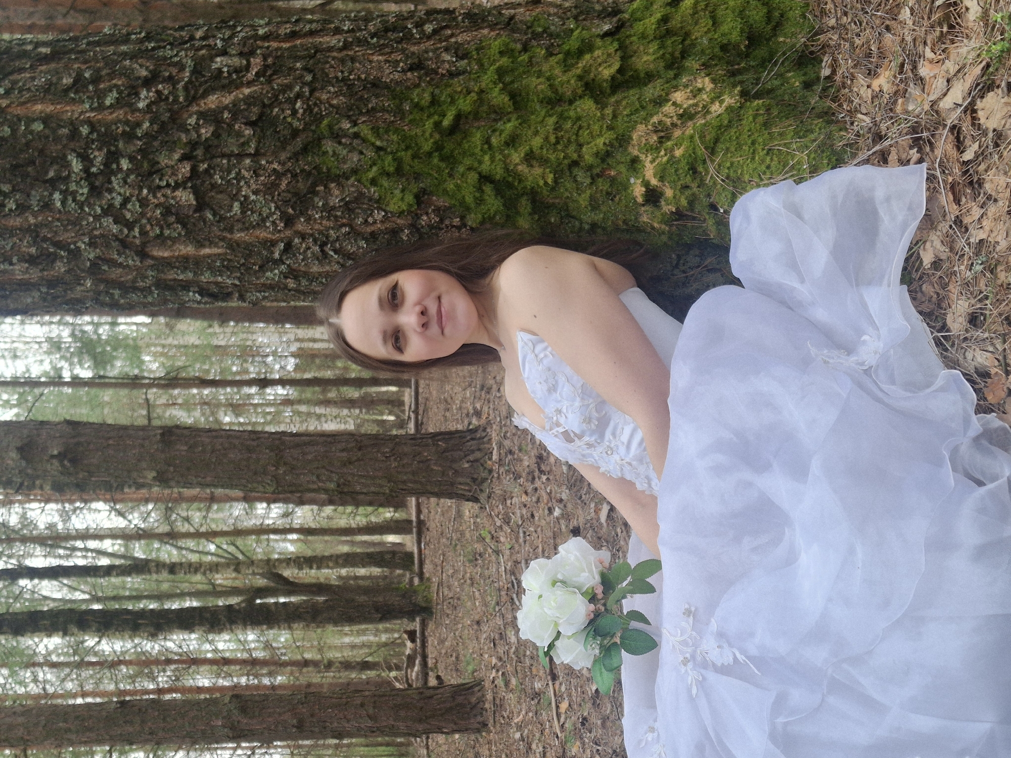 A young woman in a white wedding dress poses in a forest near Chaikovsky, Russia. She is leaning against a tree with moss on its bark and holding a bouquet of white flowers. The background includes tall pine trees and fallen leaves on the ground. The lighting is natural, suggesting an outdoor setting on a possibly overcast day.