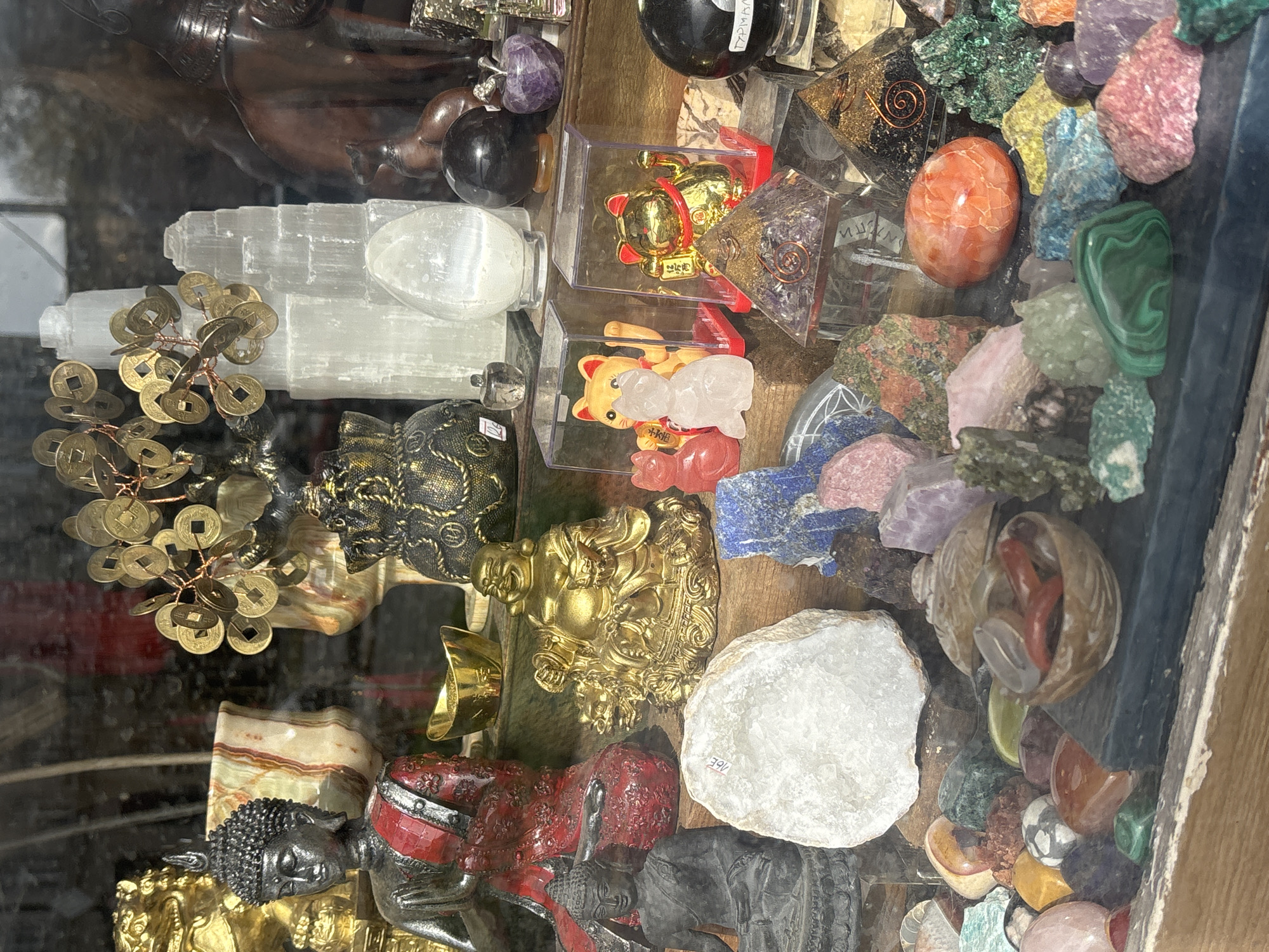 The image shows a display of various objects in a shop in Varna, Bulgaria. Prominently featured are several Buddha statues in different colors (gold, black, red), a money tree with metal coins, and selenite crystal bars. There are also figures of cats in plastic boxes, various crystal pyramids, and tumbled stones of different colors and textures. The overall impression is of a curio or souvenir shop with items related to spirituality, luck, or decoration.