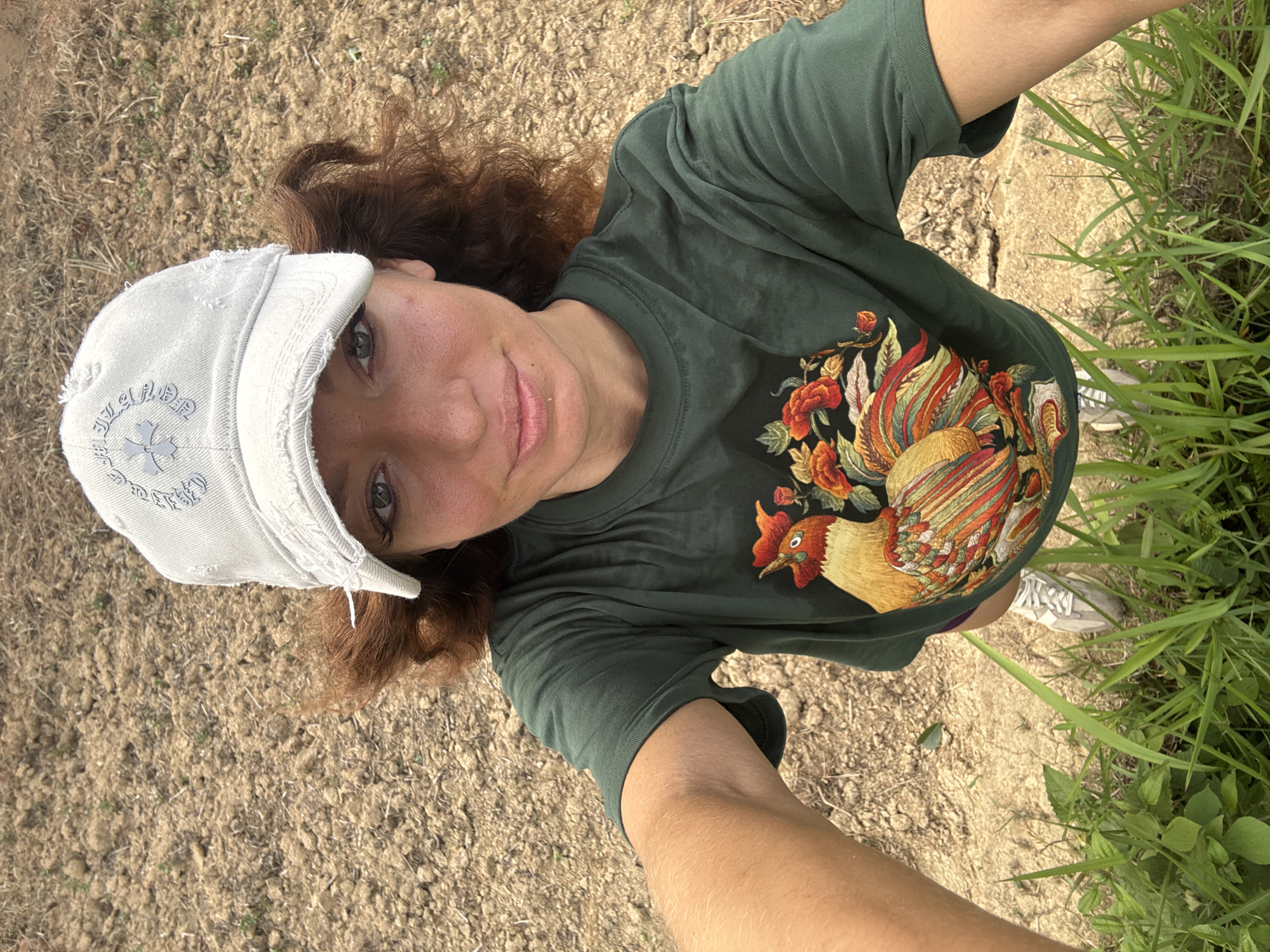 A medium shot, taken from a high angle, shows a woman outdoors in Pờ Ly Ngài Commune, Vietnam. She is wearing a distressed white baseball cap, a dark green t-shirt with a rooster and floral design on the front, and white sneakers. She has fair skin, auburn hair, and appears to be holding the camera at arm's length for a selfie. The background consists of dirt ground on the left and green grass on the right.