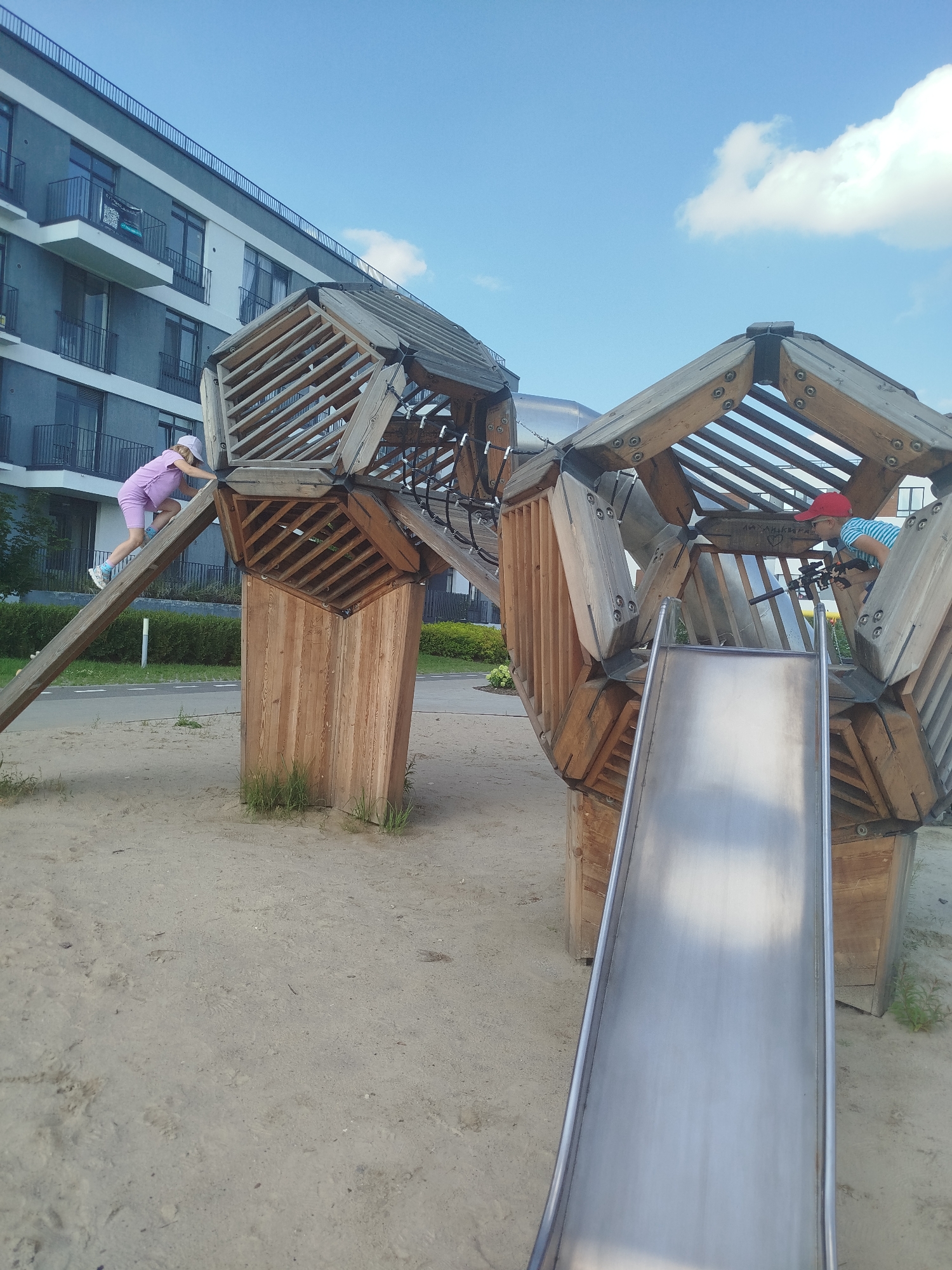 An eye-level, medium shot captures a modern, hexagonal wooden playground structure situated in a sandy area. To the left of the structure, a young child wearing a pink outfit and a white hat is climbing up a wooden ramp. In the background, a multi-story residential building with dark-tinted windows and balconies is visible under a bright blue sky with scattered white clouds. The playground features a metal slide on the right side.