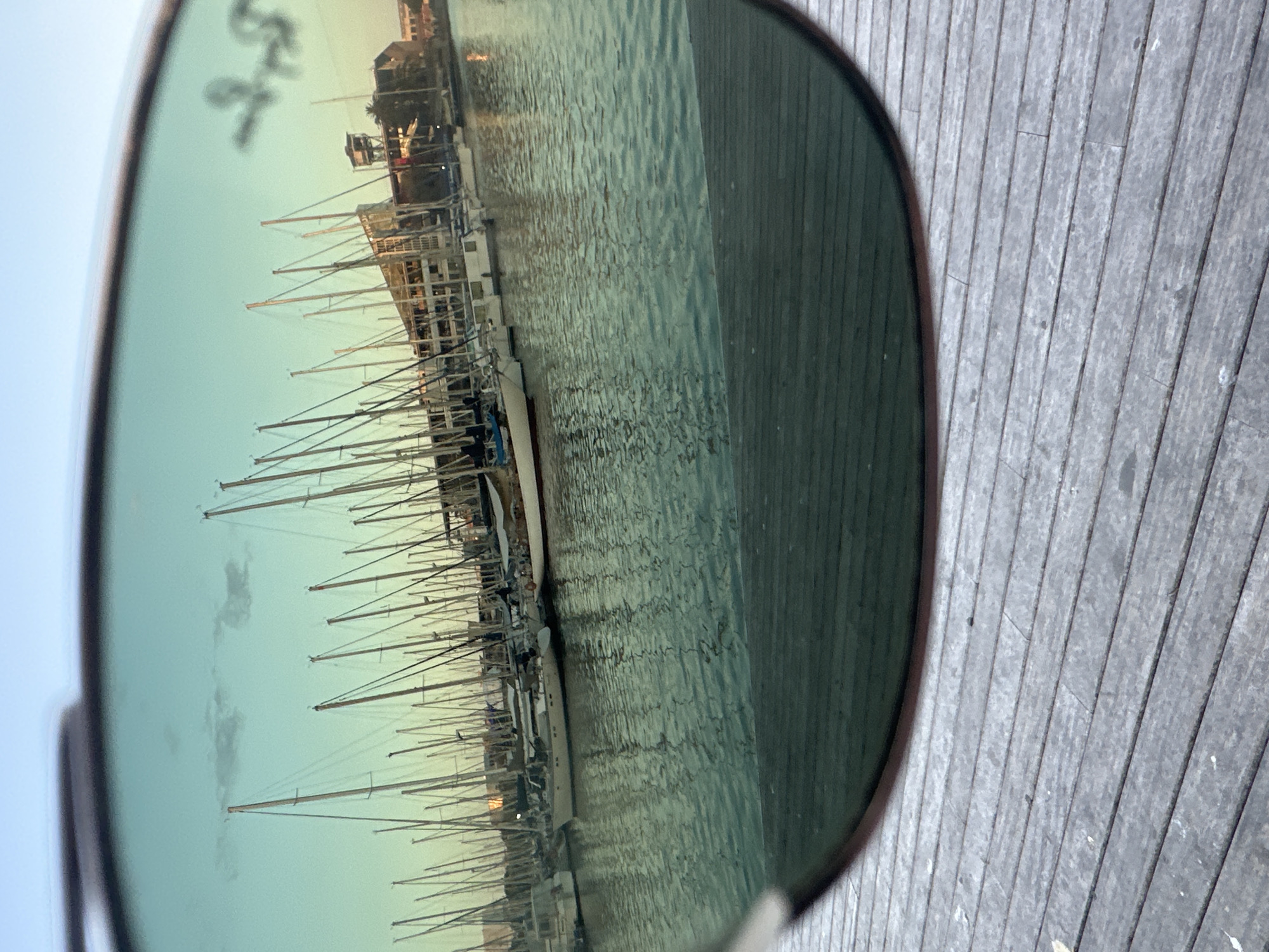The image is a close-up view of sunglasses reflecting a harbor filled with sailboats. The sunglasses are positioned in the foreground, with their dark frames visible. The reflection shows a clear sky with a hint of clouds, and numerous sailboats docked in the water. Some of the boats have white hulls and tall masts. In the background of the reflection, there is a building with multiple windows and a tower structure, typical of a waterfront area. The water is calm with ripples and reflects the sky and the boats. The original image is taken from a wooden deck, which is visible in the lower part of the frame. The scene appears to be a marina in Barcelona, Spain.