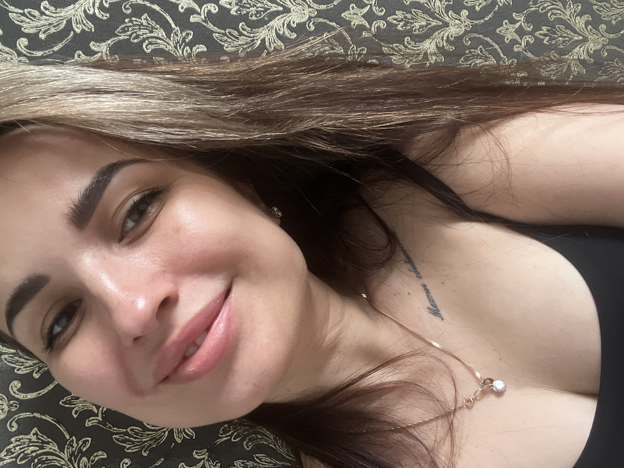 A woman with light brown and blonde hair, fair skin, and full lips is lying on a patterned sofa or cushion. She is smiling slightly and looking at the camera. She is wearing a gold necklace with a small pendant. A tattoo is visible on her chest/upper torso. The background is a patterned fabric, likely part of the sofa or cushion she is lying on. The location of the image is in the city of Unknown, Unknown.