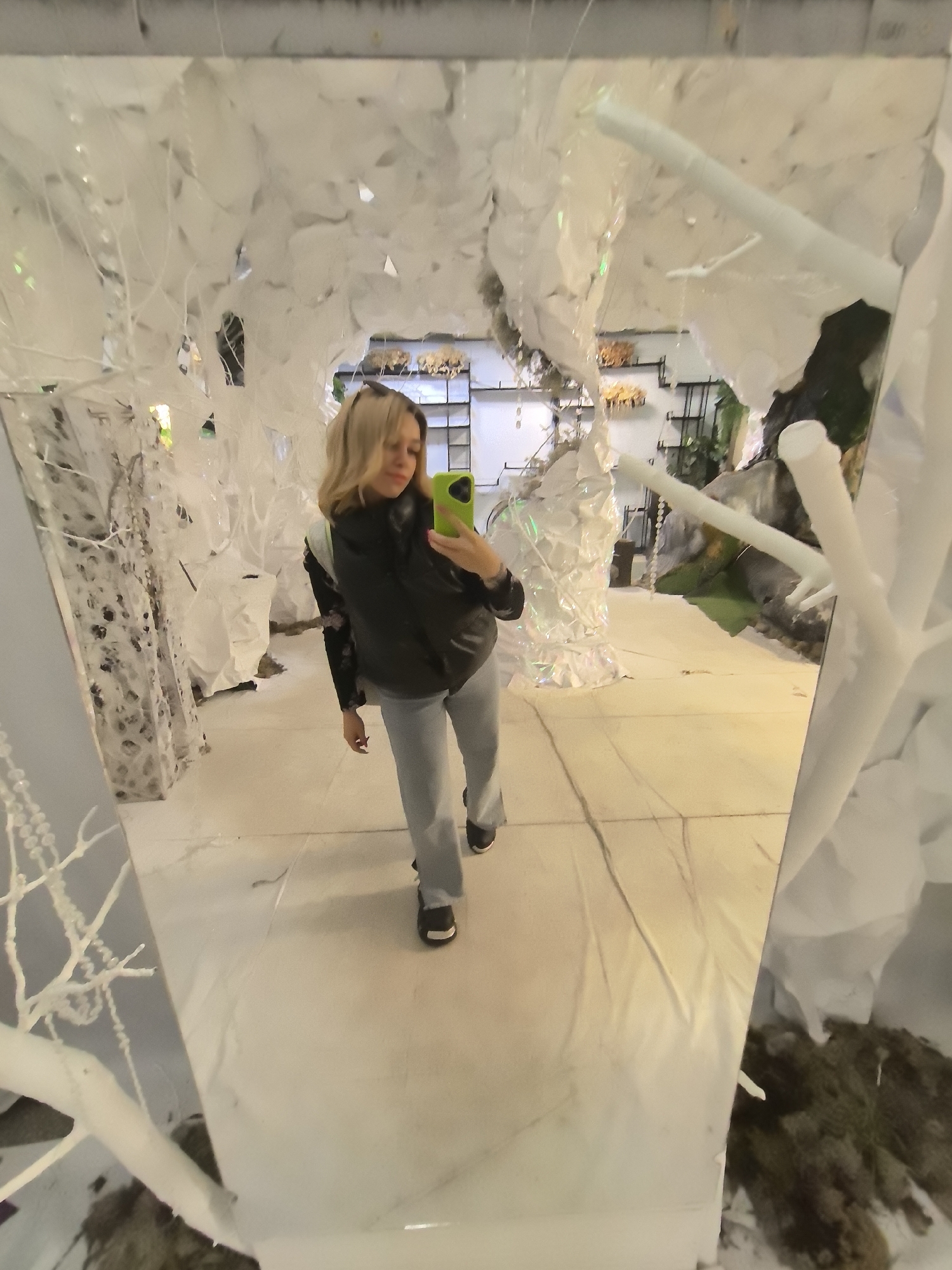 The image shows a woman taking a selfie in front of a mirror. The location appears to be an indoor space with an art installation featuring white tree-like structures and other decorative elements. The woman is wearing a black vest, a long-sleeved shirt, light blue jeans, and black shoes. She is holding a smartphone with a lime green case in her hand, using it to capture her reflection. The mirror reflects the interior space, suggesting that the installation continues beyond the immediate foreground. The city of Izhevsk, Russia, is associated with this image.