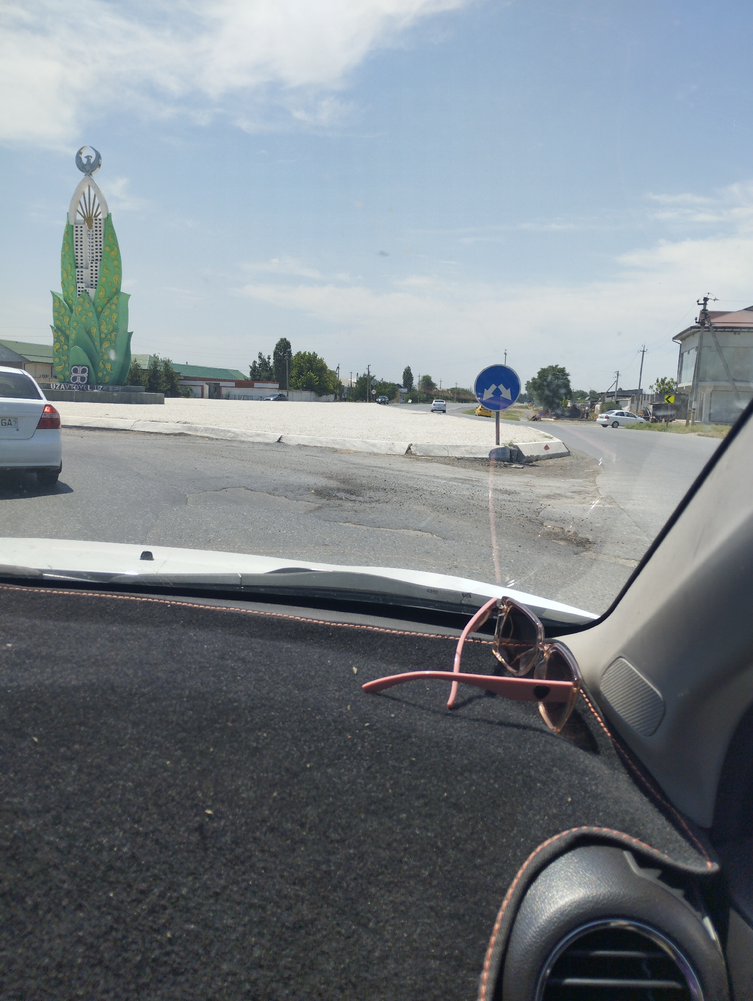 The image shows a street scene from inside a car, looking out through the windshield. A pair of pink sunglasses rests on the dashboard in the foreground. Outside, a white car is parked on the left, and a large, green monument stands in the background. The monument is tall and columnar, with a stylized bird or emblem at its apex and ornate, leaf-like green structures at its base. The monument appears to be located in a roundabout. A blue circular traffic sign with arrows indicating a roundabout is visible to the right of the monument. Buildings and trees line the background. The sky is partly cloudy. The overall environment suggests an urban or semi-urban setting, possibly in a public square or intersection in the city of Turkkishlak, Uzbekistan. The license plate of the white car is partially visible and appears to read "GA".