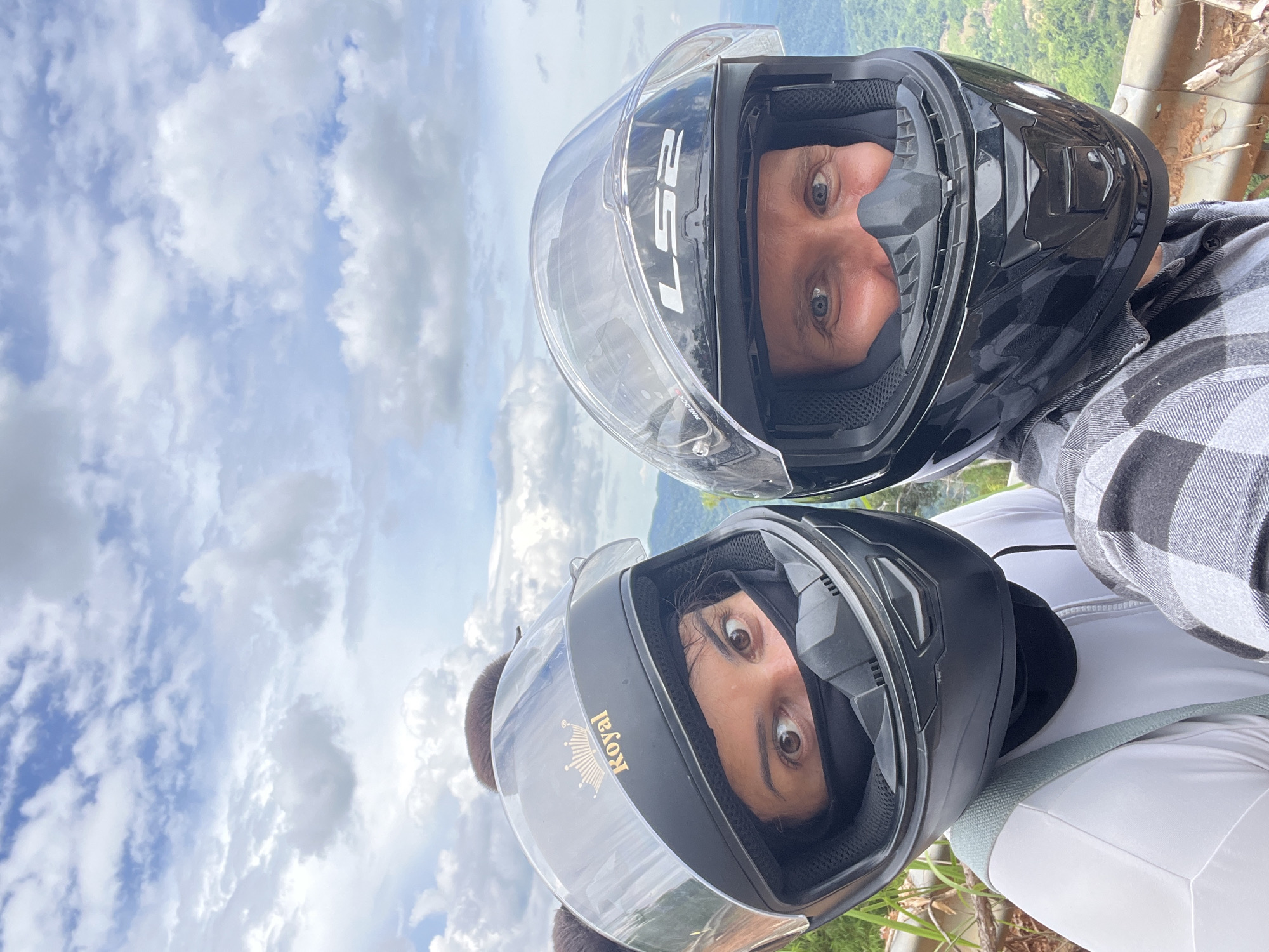 In the city of Son Dien, Vietnam, two individuals are captured in a close-up, selfie-style photograph while wearing motorcycle helmets. The person on the left, positioned slightly closer to the camera, wears a black helmet adorned with a gold crown emblem and the word "Royal" printed in gold lettering. Their eyes are visible above a black face mask, and their brown hair is styled with two pom-pom-like accessories. They are wearing a white top with a light green strap across their chest.
The person on the right wears a black helmet with a clear visor, displaying the "LS2" logo. Their blue eyes are visible, and they are wearing a black nose guard. They are dressed in a grey and black plaid shirt. Both individuals appear to be looking towards the camera.
The background of the image reveals a lush, green landscape with rolling hills and trees, indicative of a rural or mountainous area. The sky above is filled with scattered white clouds against a bright blue backdrop, suggesting daylight conditions. A blurred effect on the background hints at the subjects being in motion or the focus being intentionally on the individuals.