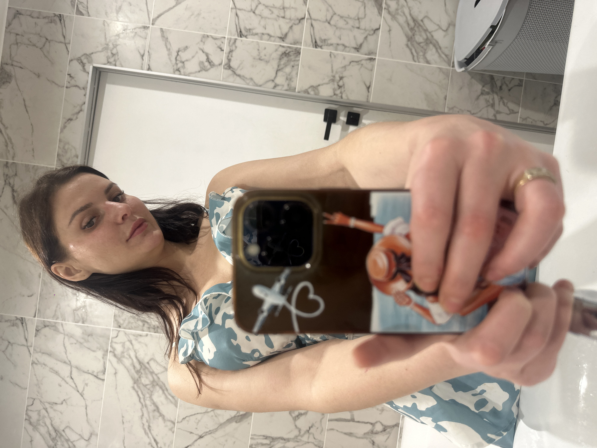A young woman takes a mirror selfie in a bathroom. She is wearing a blue and white floral dress. Her hair is dark brown and pulled back. Her left hand is holding up her phone, which has a case with images of an airplane and a person wearing a hat. Her right hand is visible, with a ring on her finger. The bathroom has marble-patterned tiles on the walls and a white sink. A gray trash can is visible in the background.