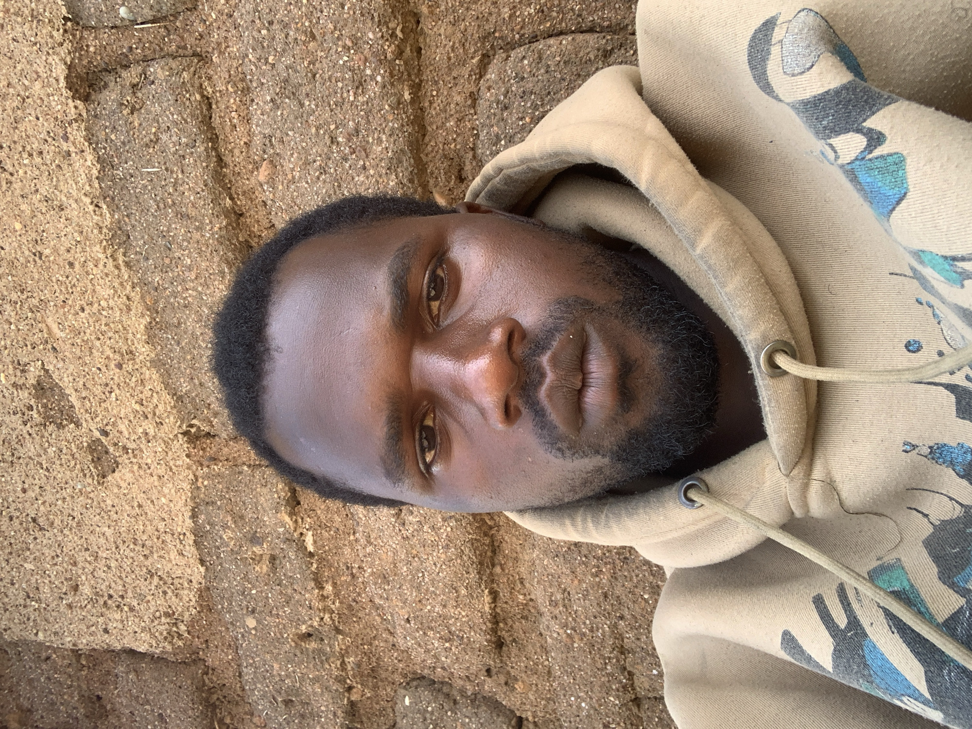 The image is a head-and-shoulders selfie of a Black man with dark skin and short, black hair, and a beard. He is wearing a light brown hooded sweatshirt with drawstrings. The background consists of a rough, textured wall made of brown bricks or mud. The man's face is centered in the frame, looking directly at the camera. The lighting appears natural and somewhat harsh, casting subtle shadows on his face. The setting is an outdoor environment. Based on the provided information, this scene is located in Buga da Gindi, Nigeria.