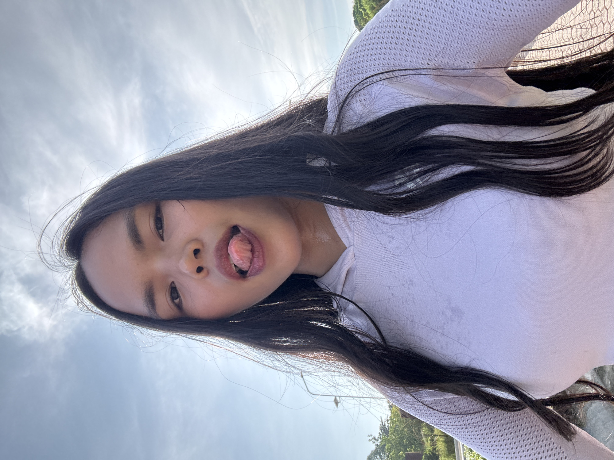 A young woman with long, dark hair is pictured outdoors, with a bright sky and scattered clouds behind her. She is wearing a white, long-sleeved top with a textured pattern on the sleeves and a smooth front. She is looking towards the camera with her tongue sticking out. The lighting suggests it is daytime. The location is described as Davao City, Philippines.