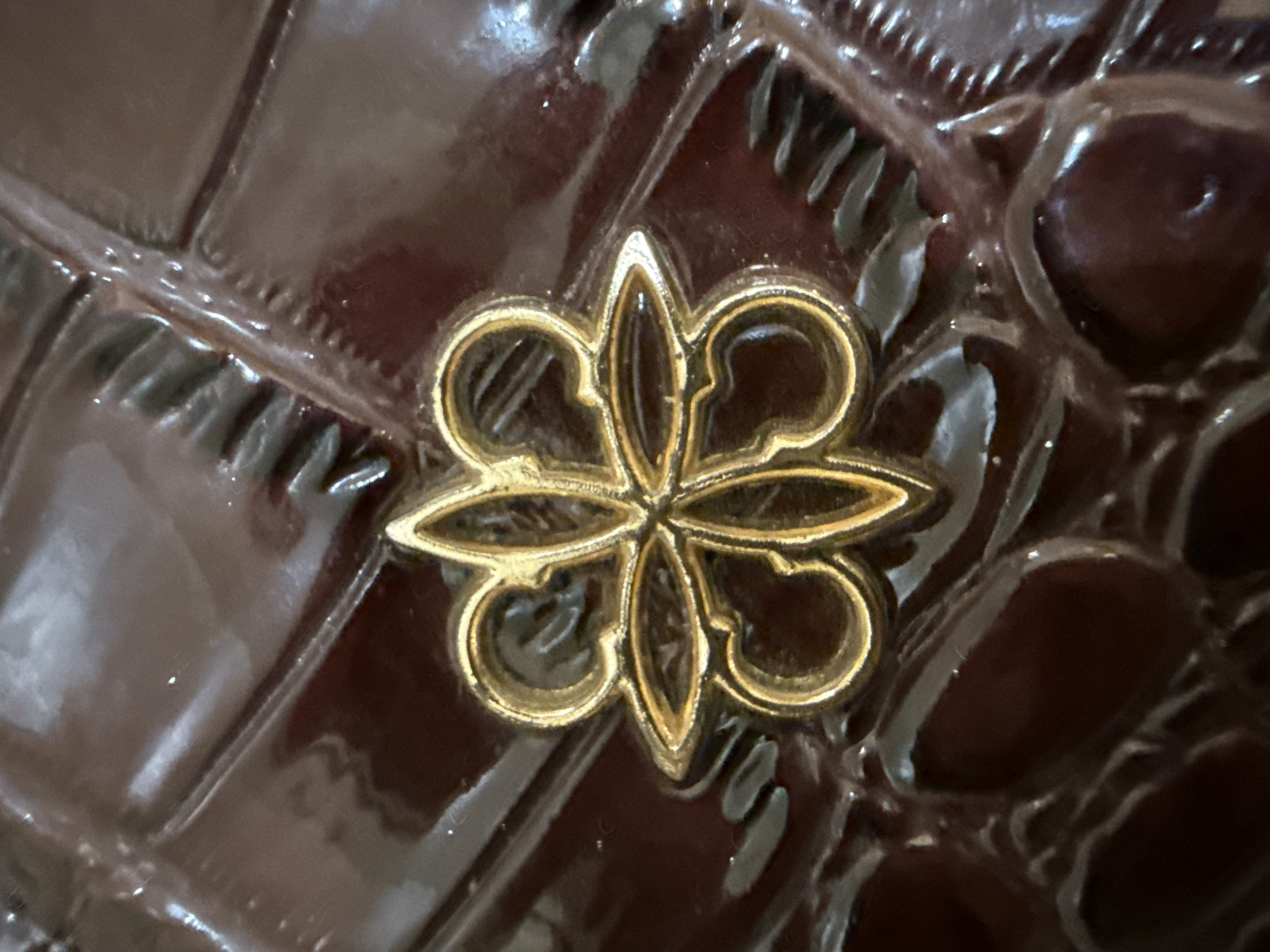 The image is a close-up shot of a gold-colored emblem, possibly a logo or decorative piece, placed on a dark, glossy, textured surface. The emblem is intricate, appearing to be made of four overlapping, stylized petal-like shapes with curved stems. The surface it rests on has a diamond-shaped pattern, with the glossy material creating highlights and shadows that emphasize the texture. There are no people, actions, or interactions visible. The specific location in the city of Lhokseumawe, Indonesia, cannot be determined from the provided image.