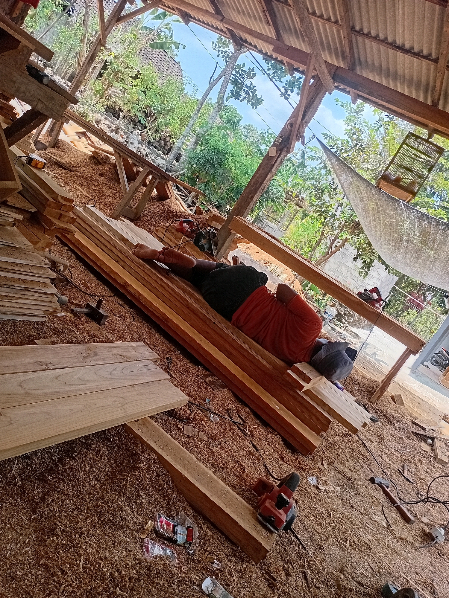 The image depicts an outdoor woodworking area in Playen, Indonesia. A person, wearing an orange shirt and dark shorts, is lying down on a stack of lumber, appearing to rest. The ground is covered in sawdust, indicating active woodworking. Several pieces of lumber are stacked around the person and on a workbench. A power planer and a hammer are visible near the lumber, suggesting ongoing or recently completed work. The scene is situated under a rustic, open-sided structure with a corrugated metal roof. In the background, lush green vegetation and trees are visible, characteristic of the tropical environment of Indonesia. The overall impression is one of a practical, working space amidst a natural setting.