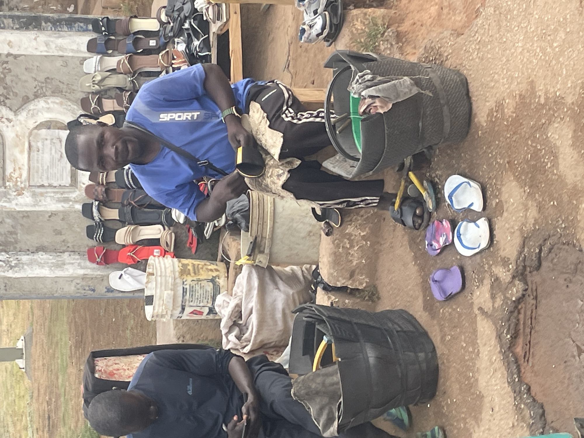The image shows two men seated in an outdoor setting, likely a marketplace. One man wears a blue "Sport Sport" t-shirt and dark pants with a white stripe. He sits amidst a display of sandals and footwear, holding a dark object. Near him are flip-flops, a black container with items inside, and a bucket. The other man, wearing a dark long-sleeved shirt, is seated near another similar black container. The ground is textured, potentially dirt or concrete. The city is Jos, Nigeria.
