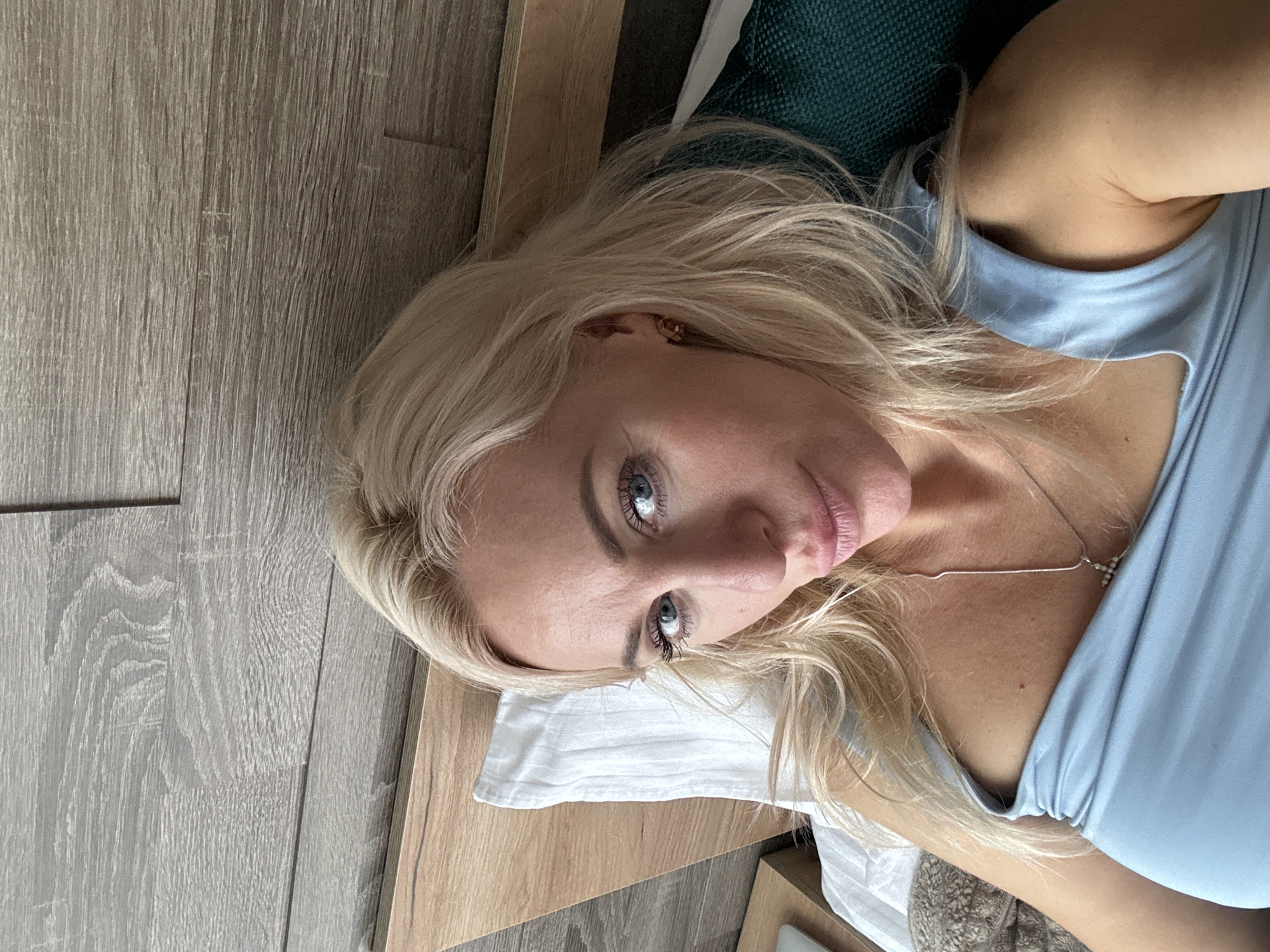 A close-up, high-angle shot shows a woman with blonde hair and blue eyes lying on a bed in Starye Chernitsy, Russia. She is wearing a light blue top and a silver necklace. Her face is turned towards the camera, and she has a neutral expression. The background consists of a wooden floor and a dark green headboard. The lighting suggests it is daytime. There is no visible text in the image.