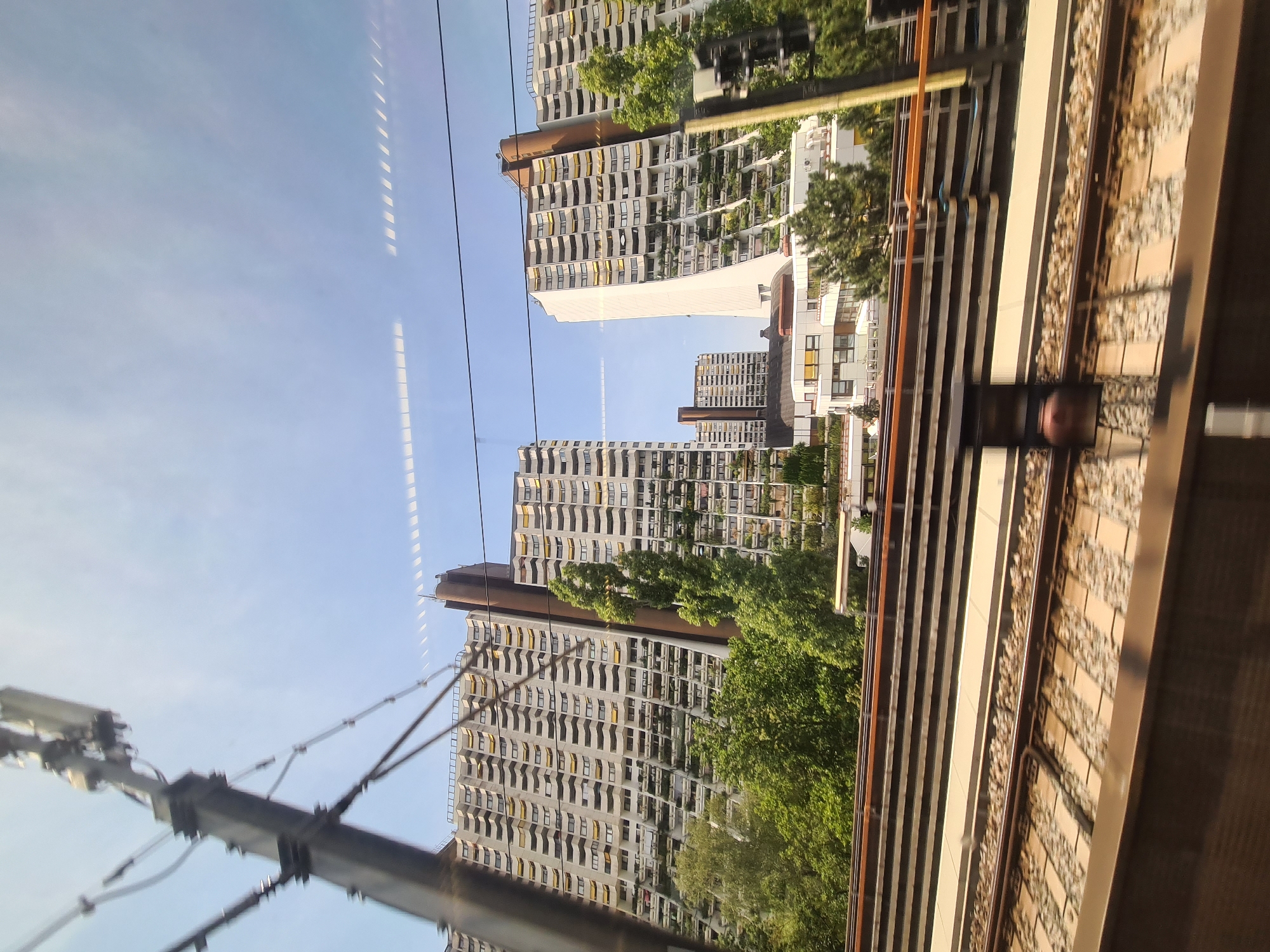 The image depicts a view from a moving train in Vienna, Austria, focusing on a distinctive architectural complex and the railway tracks in the foreground. The prominent structures are large residential buildings with numerous balconies adorned with yellow awnings and greenery, suggesting a well-populated urban area. One building has a striking curved facade. In the background, a blue sky with scattered clouds is visible. The foreground shows multiple sets of railway tracks, with ballast and sleepers clearly discernible, indicating active train lines. A tall, dark pole with electrical equipment and wires is present on the left side of the frame, partially obscuring the view of the buildings. The overall impression is one of an urban landscape viewed from a transportation route.