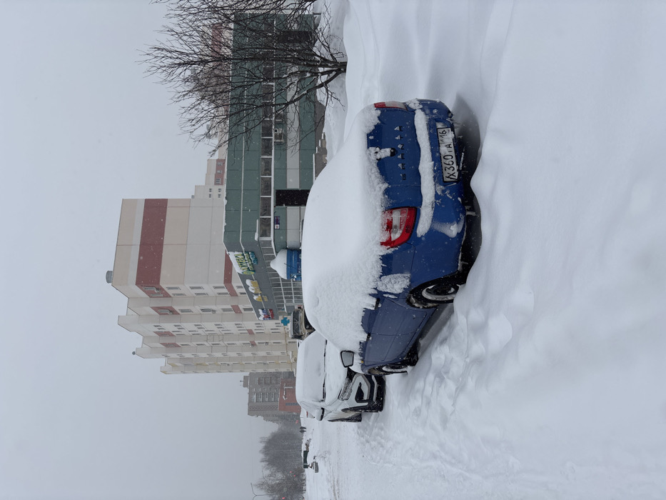 The image depicts a winter street scene in Nizhnekamsk, Russia, under overcast skies with active light snowfall. In the foreground and mid-ground, two vehicles are parked parallel to a snow-covered bank. The closest vehicle is a blue Lada Granta sedan, heavily covered in fresh snow on its roof, trunk, and rear window, with Russian license plate X360TA 116 visible. Behind it, a white SUV is also substantially obscured by snow. Further back, a dark-colored vehicle is partially visible.
In the background, a multi-story apartment building, characterized by white, red, and beige horizontal bands, stands prominently. In front of this building, a smaller, single-story commercial structure with a teal-green facade features multiple windows and a main entrance. Signage on this building clearly displays "АПТЕКА" (Pharmacy). A blue, dome-shaped object, partially covered in snow, is situated on the ground near the commercial building.
On the far left, a snow-covered path or street extends into the distance, where two indistinct human figures are visible walking. The entire visible ground surface is blanketed in a thick layer of snow, with faint tracks indicating prior passage. Bare deciduous trees are present on the right side of the frame.