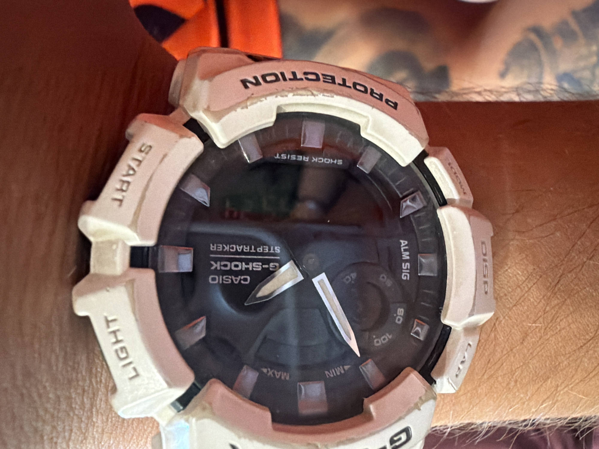The image shows a close-up of a white Casio G-Shock Steptracker watch on a person's wrist. The watch face is black with silver hands and markings. The words "LIGHT", "START", "SHOCK RESIST", and "PROTECTION" are visible on the bezel and surrounding case. Some of the watch's buttons and indicators are also visible. The background is out of focus, but appears to be an outdoor setting with some skin and clothing visible. The image is taken from a slightly elevated angle, looking down at the watch.
