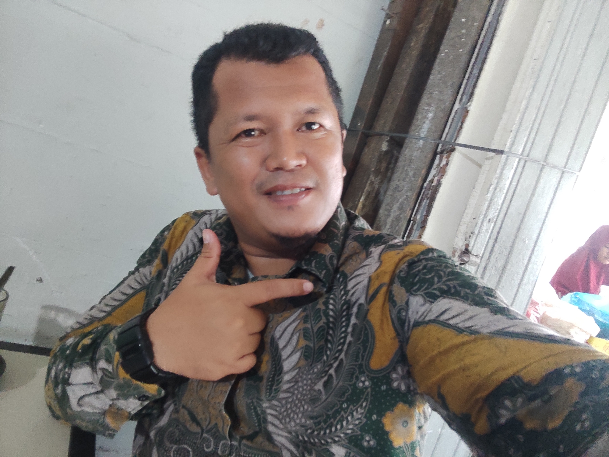 A man poses for a selfie indoors. He has dark hair and skin, and is wearing a patterned green and yellow shirt. He is pointing at himself with his right hand. He has a black smartwatch on his left wrist. Behind him are a white wall and some metal structures. Another person wearing a red hijab is partially visible in the background. The location is Muliengmanyang, Indonesia.