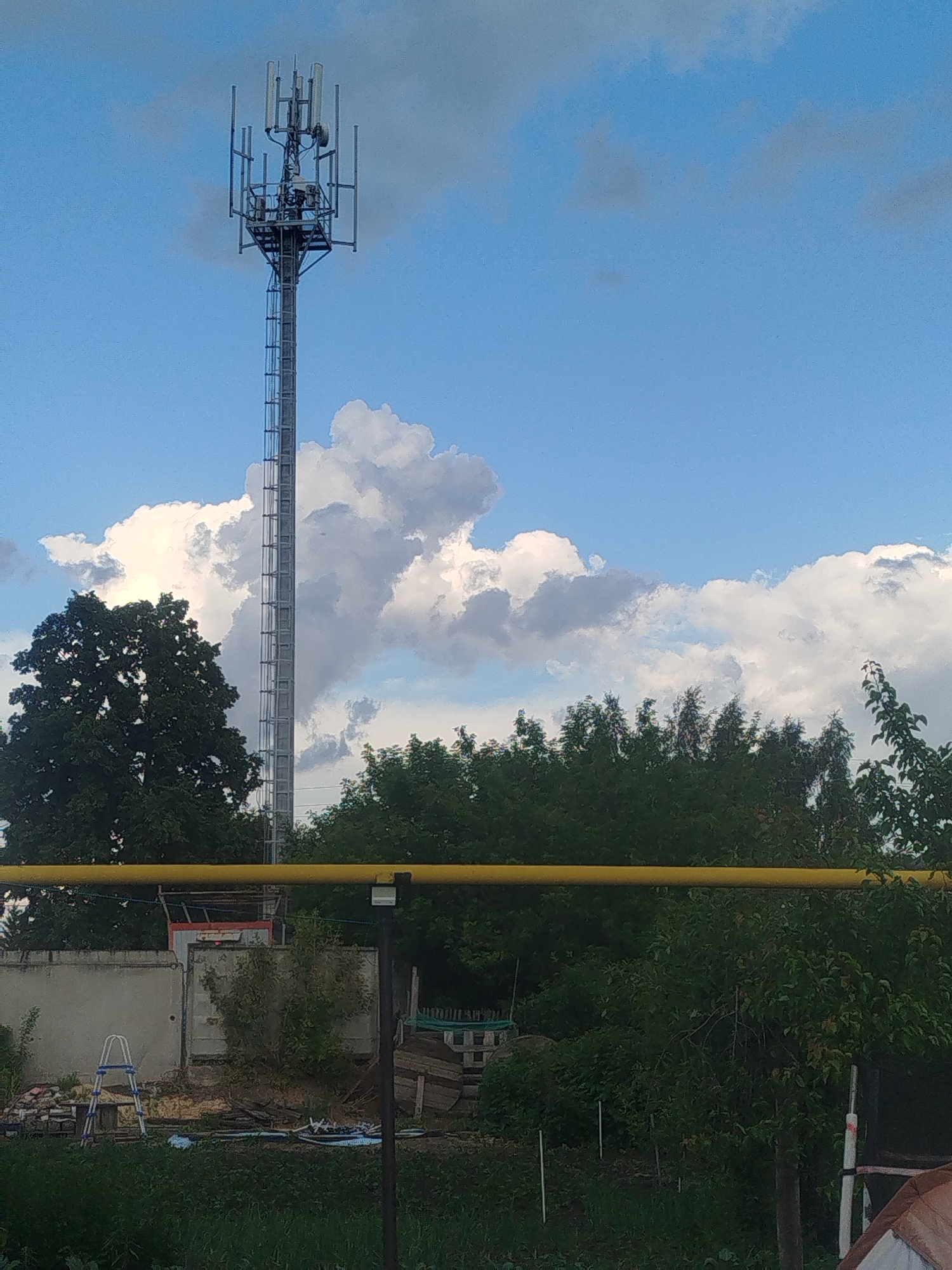 The image shows a cell tower against a blue sky with white and gray clouds. The tower is tall and metallic, with multiple antennas at the top. In the foreground, there is a yellow pipe running horizontally across the frame. Below the pipe, there is a line of trees and vegetation. To the left, there is a concrete wall with a ladder leaning against it, and some debris. The scene appears to be in a semi-rural or industrial area. The location is stated to be Krasny Gulyai, Russia.